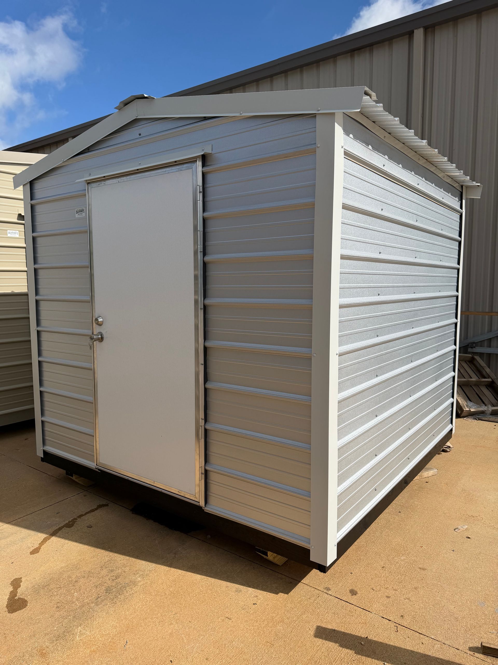 Small silver corrugated metal shed with a white door on a sunny concrete lot