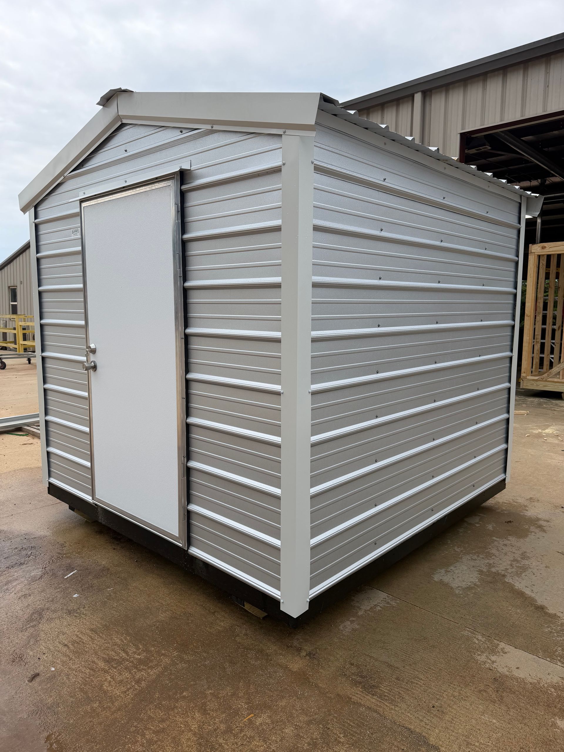 Small silver metal storage shed with a white door on a wet industrial lot