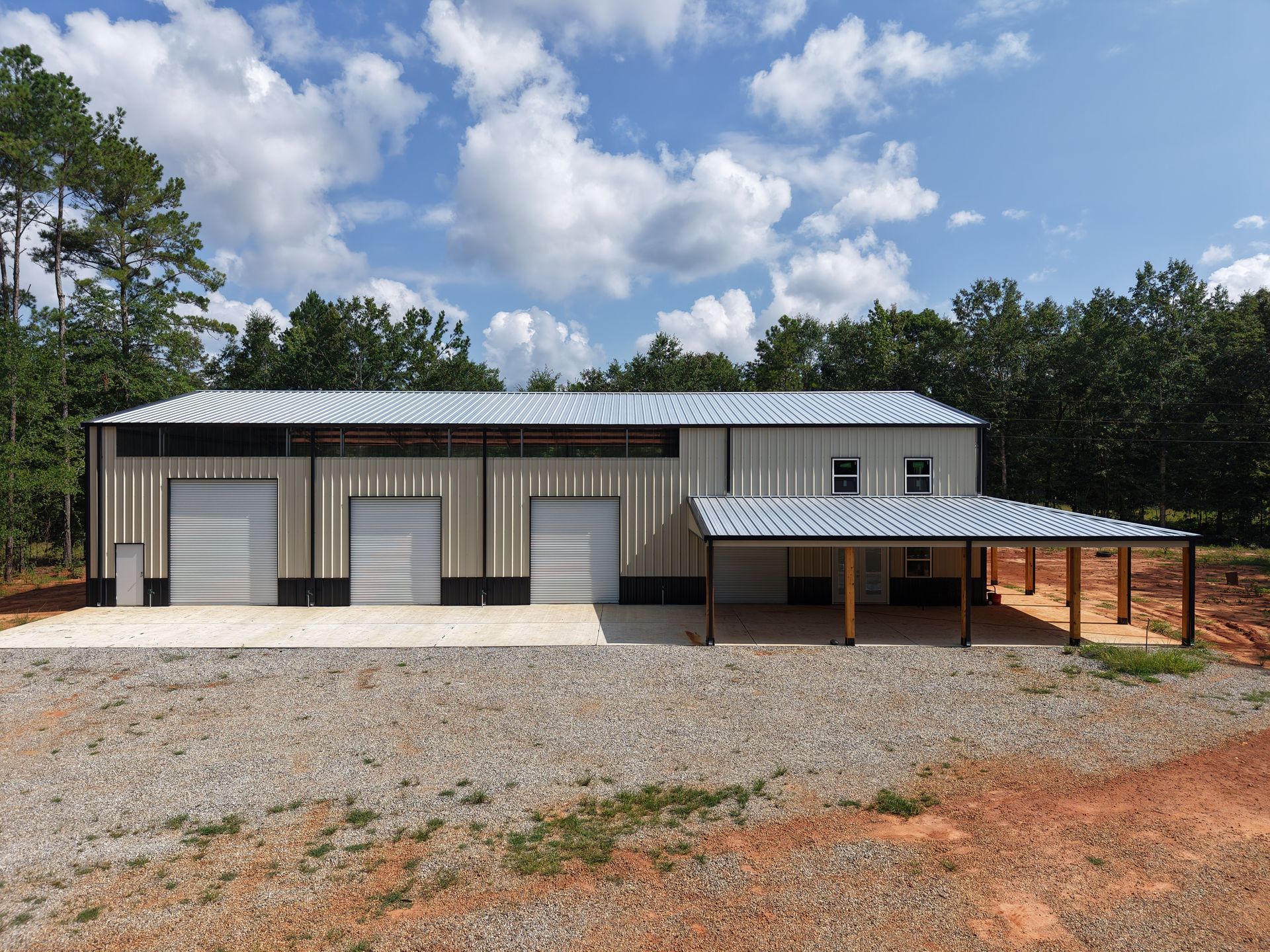 Metal building with three garage doors and a covered porch. Gray, beige, and brown color scheme. Gravel driveway.
