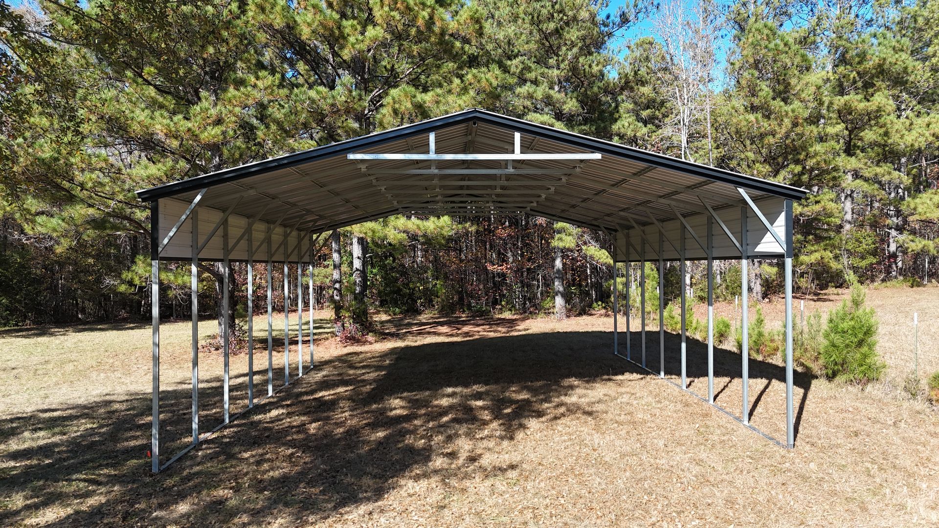 Metal carport with a peaked roof, standing in a clearing surrounded by trees.