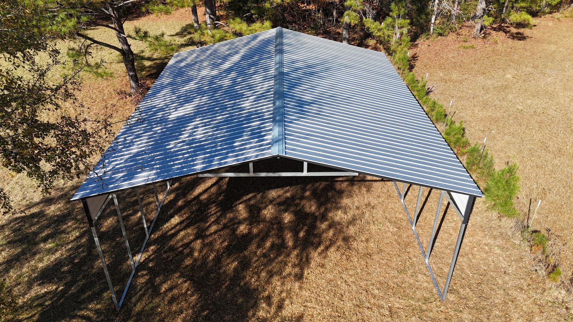 Carport with corrugated metal roof on a grassy area, surrounded by trees.