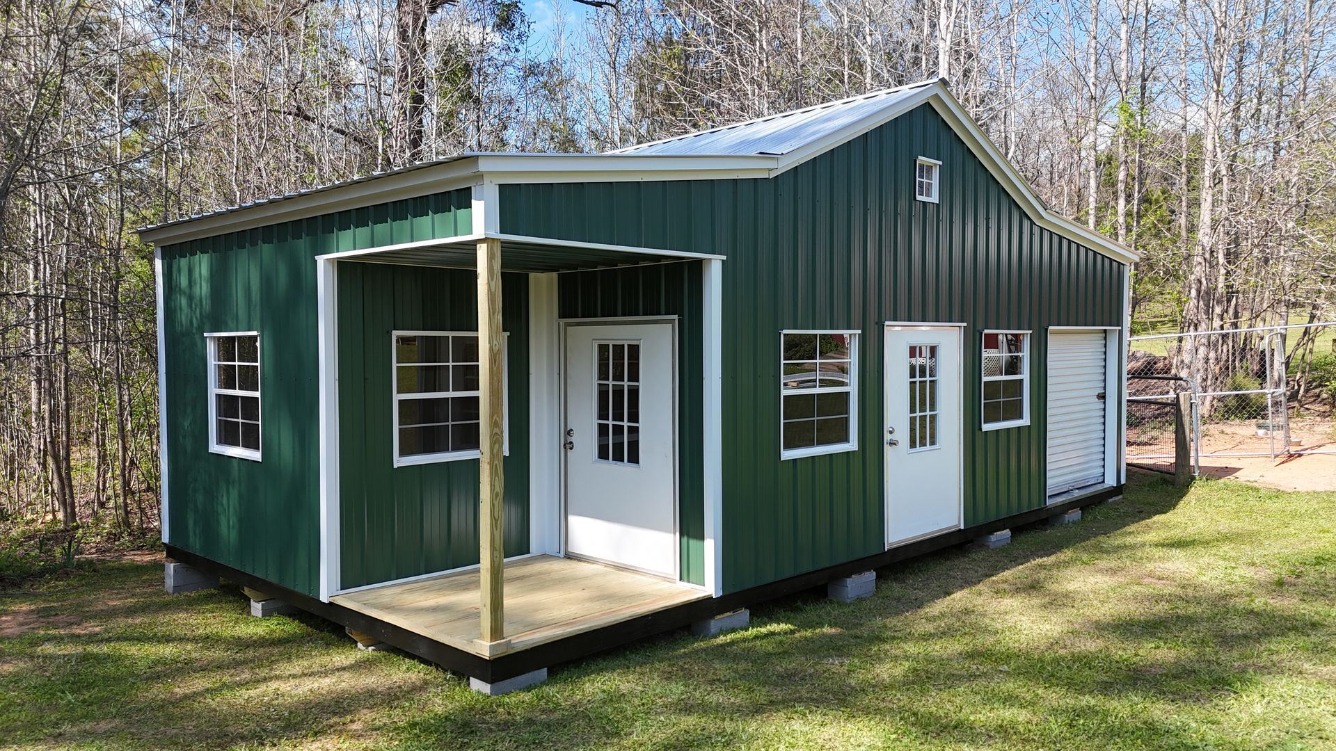 A green, metal-sided shed with a front porch, two white doors, several windows, and a roll-up door in a wooded setting.