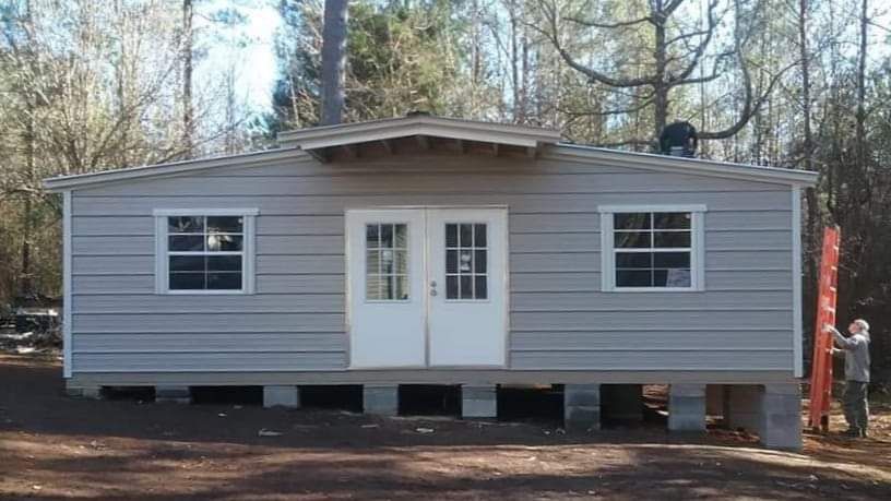 Gray shed-like building with double doors and windows on concrete blocks; person near a ladder on the right.
