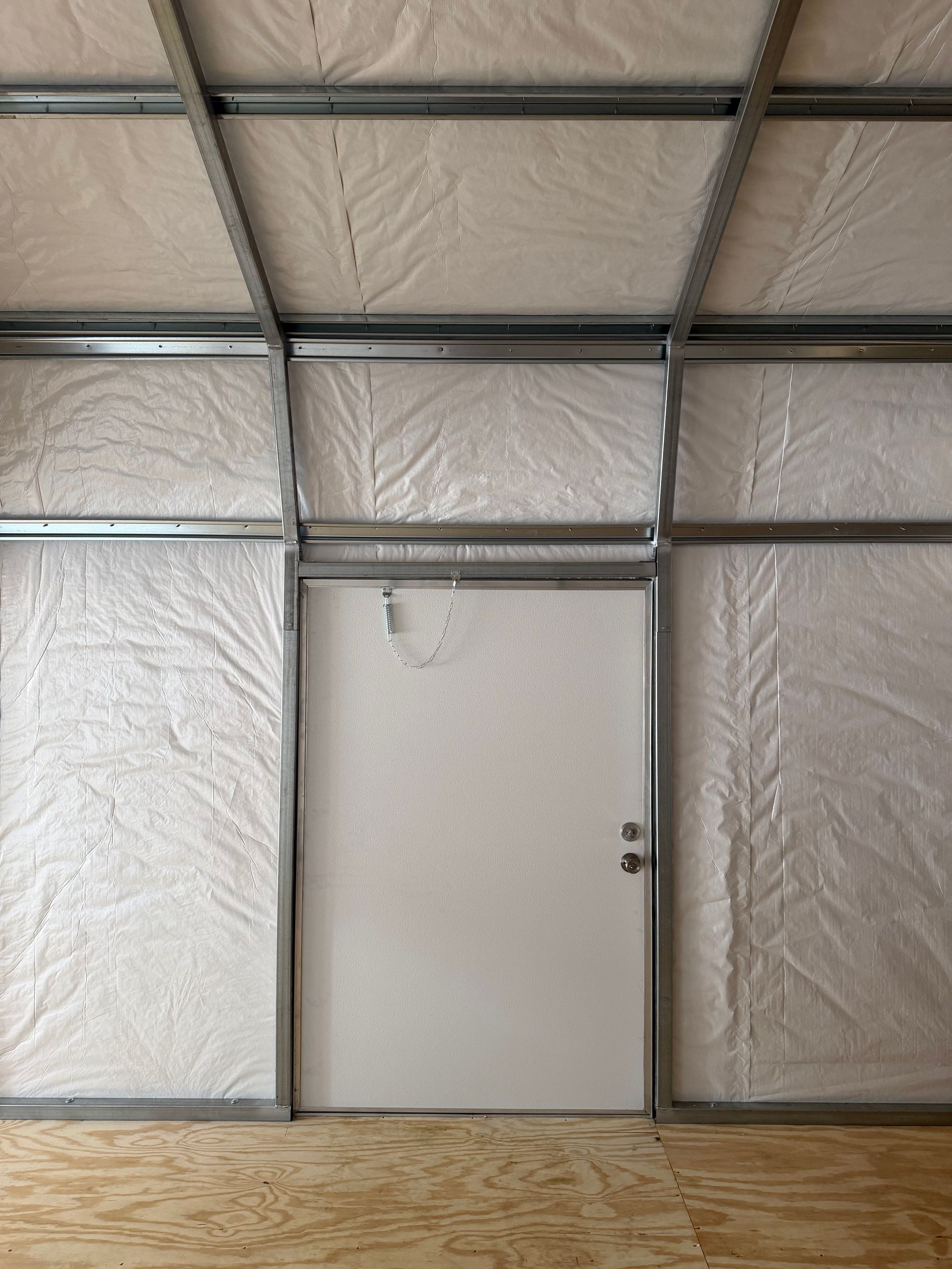 Interior wall with exposed metal framing, insulated panels, and a closed door over a plywood floor.