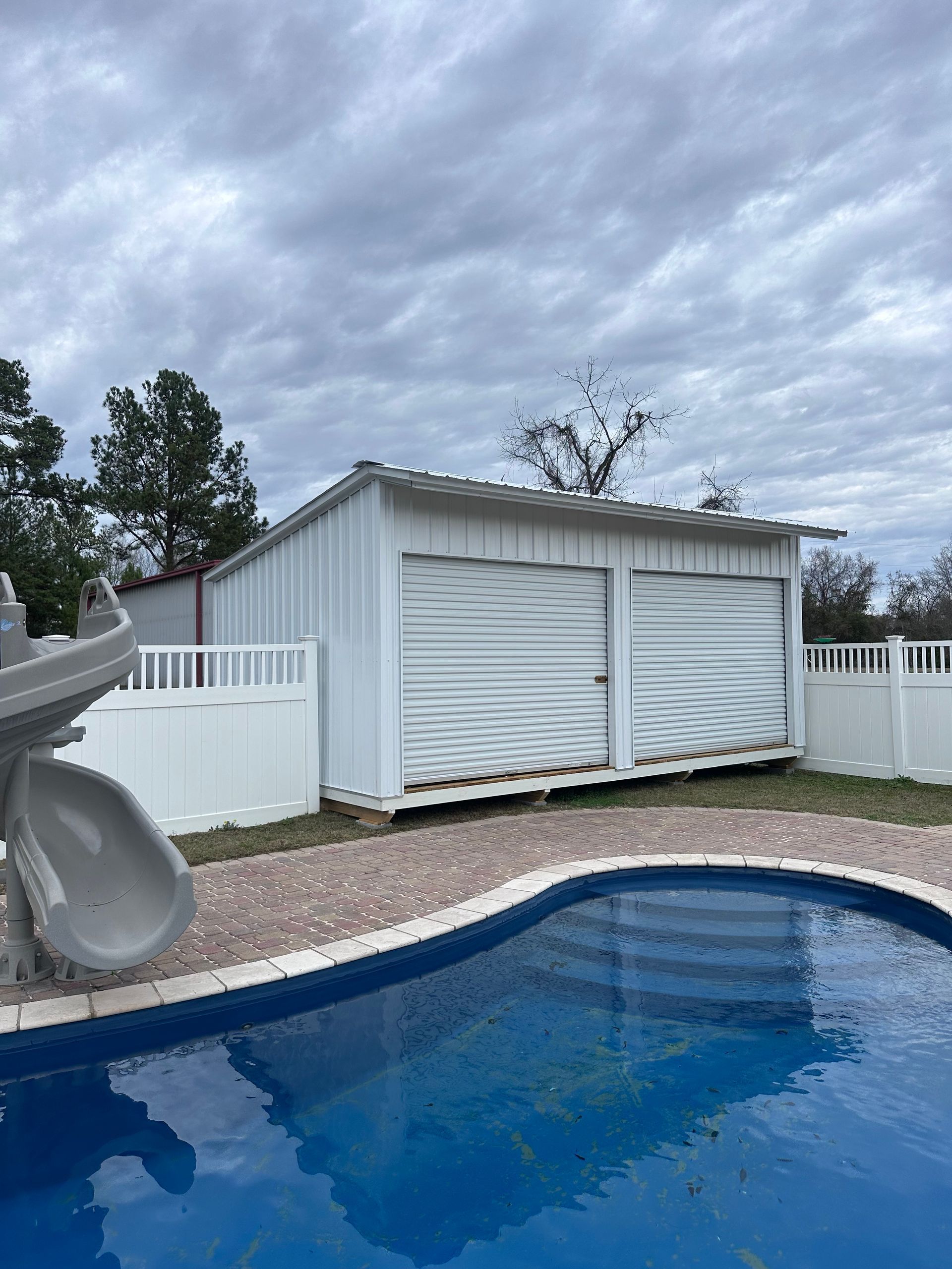 White shed with two roll-up doors, next to a pool with a slide, under a cloudy sky.