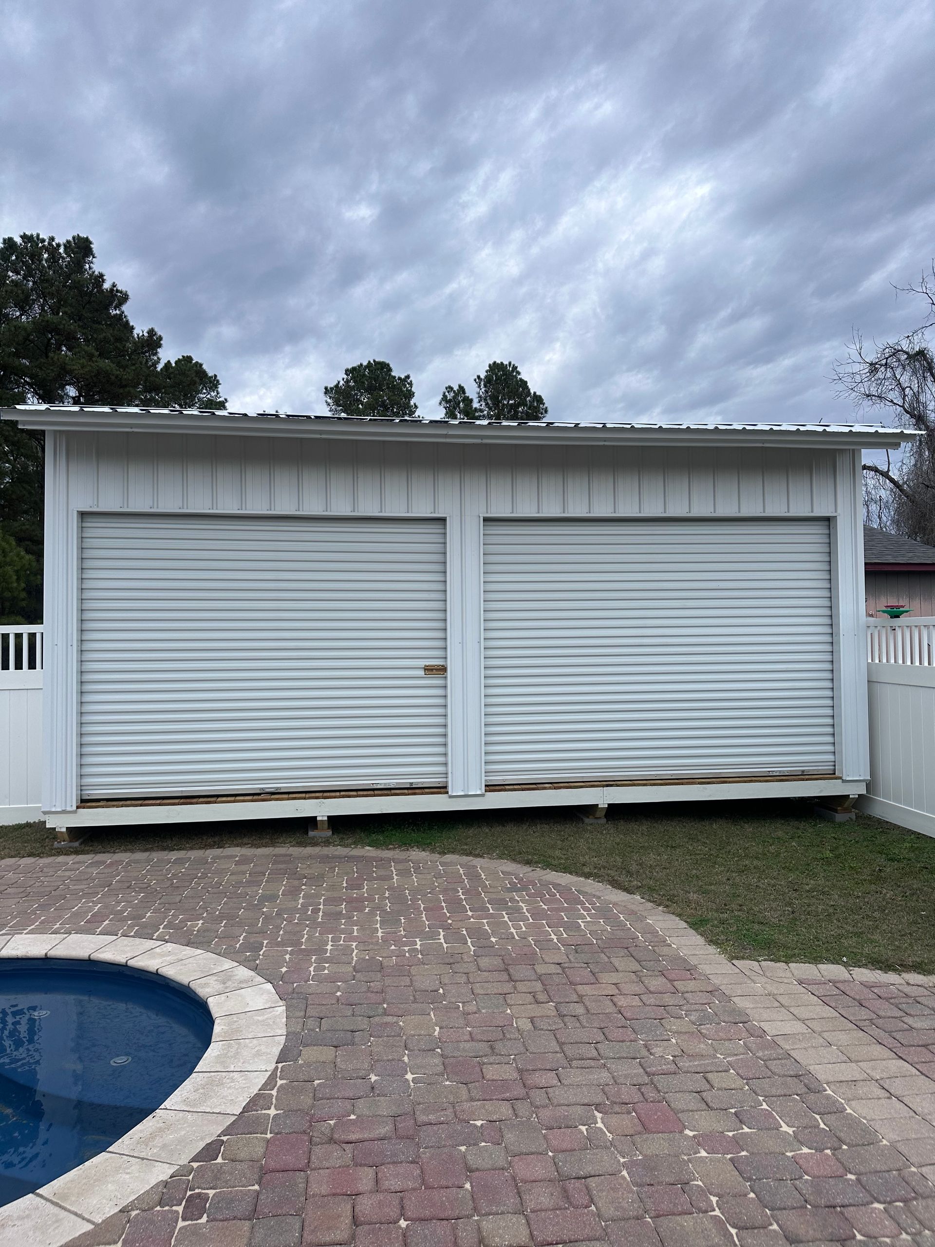 White shed with two closed garage doors, next to a pool and brick patio, under a cloudy sky.