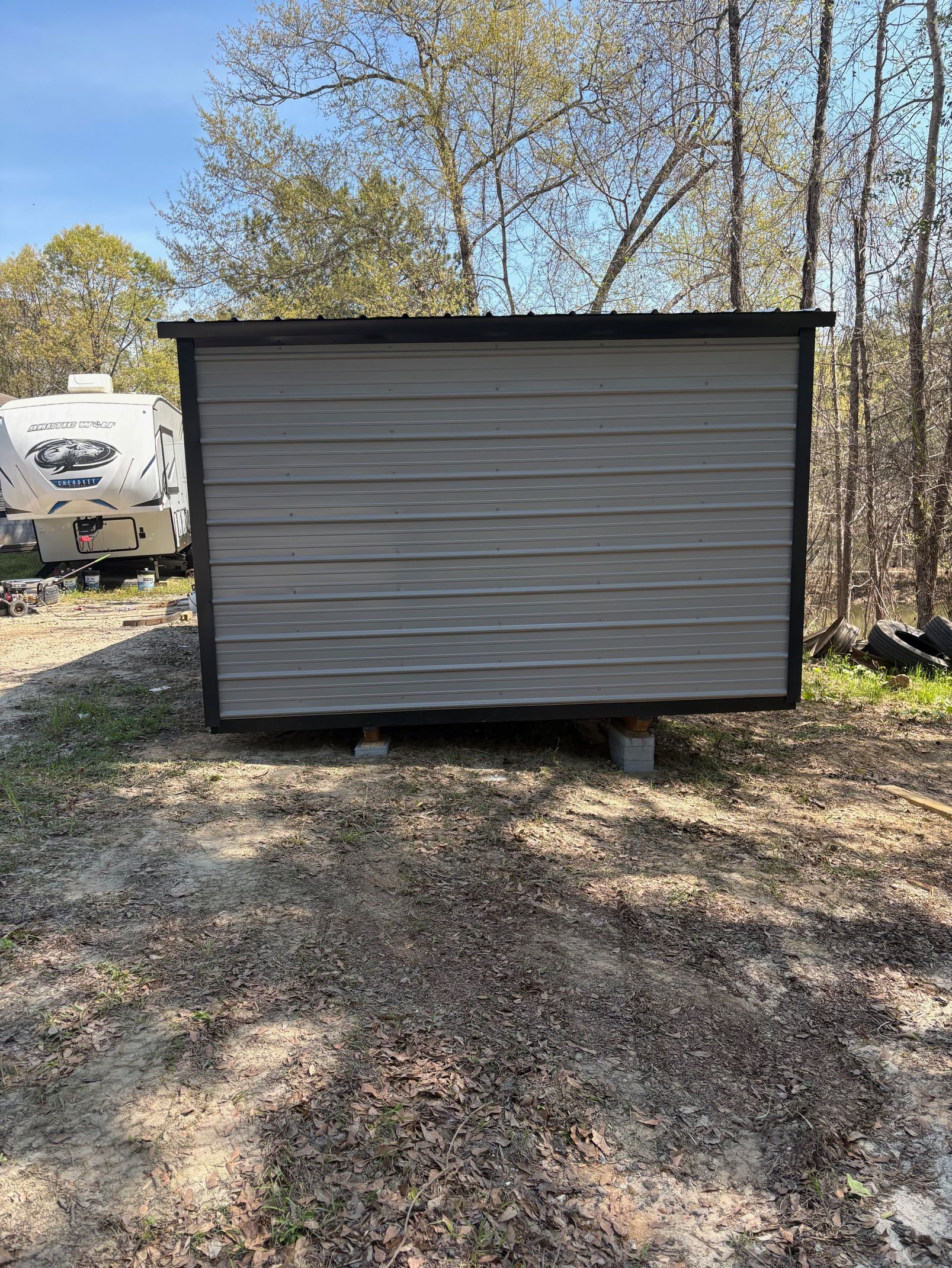 A rectangular storage shed with grey horizontal siding and black trim, resting on concrete blocks in a wooded area.