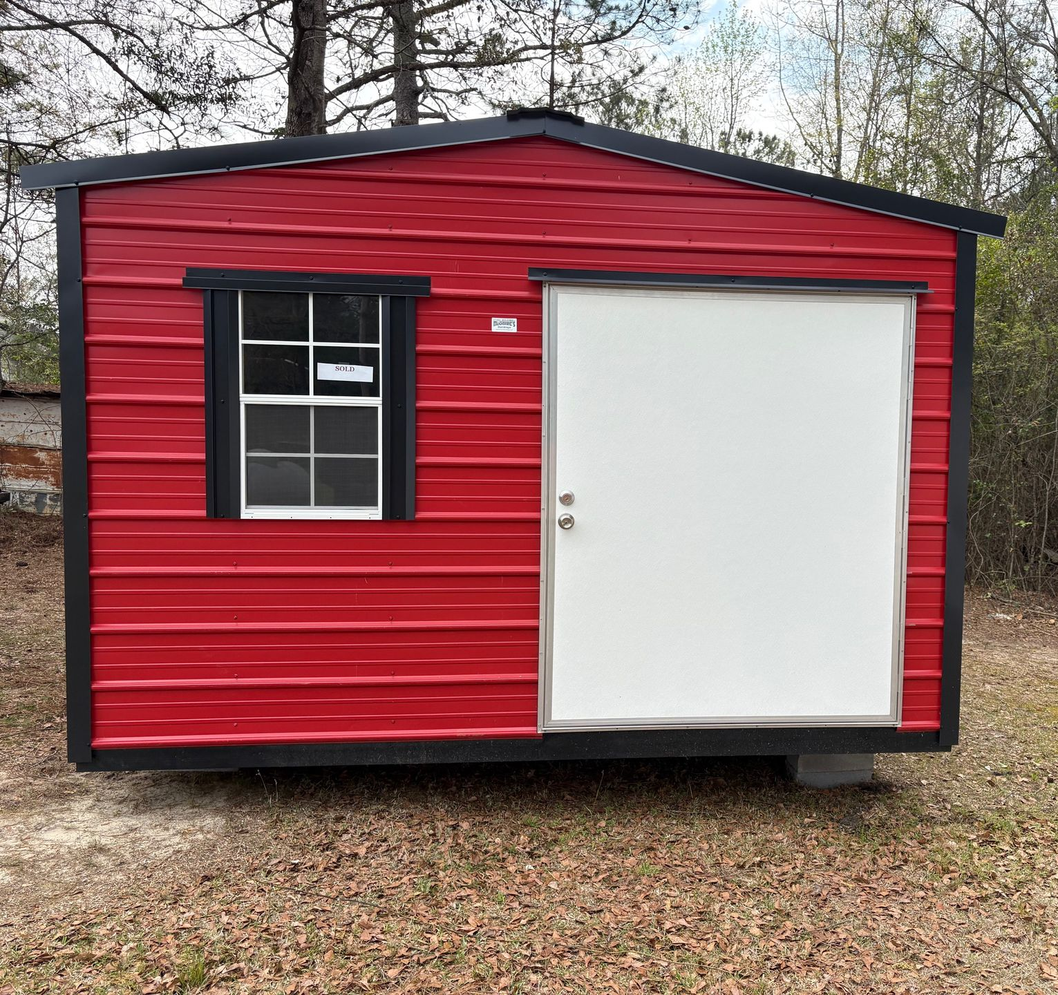 A red metal storage shed with a white door and a single window with black trim, situated in a wooded outdoor setting.