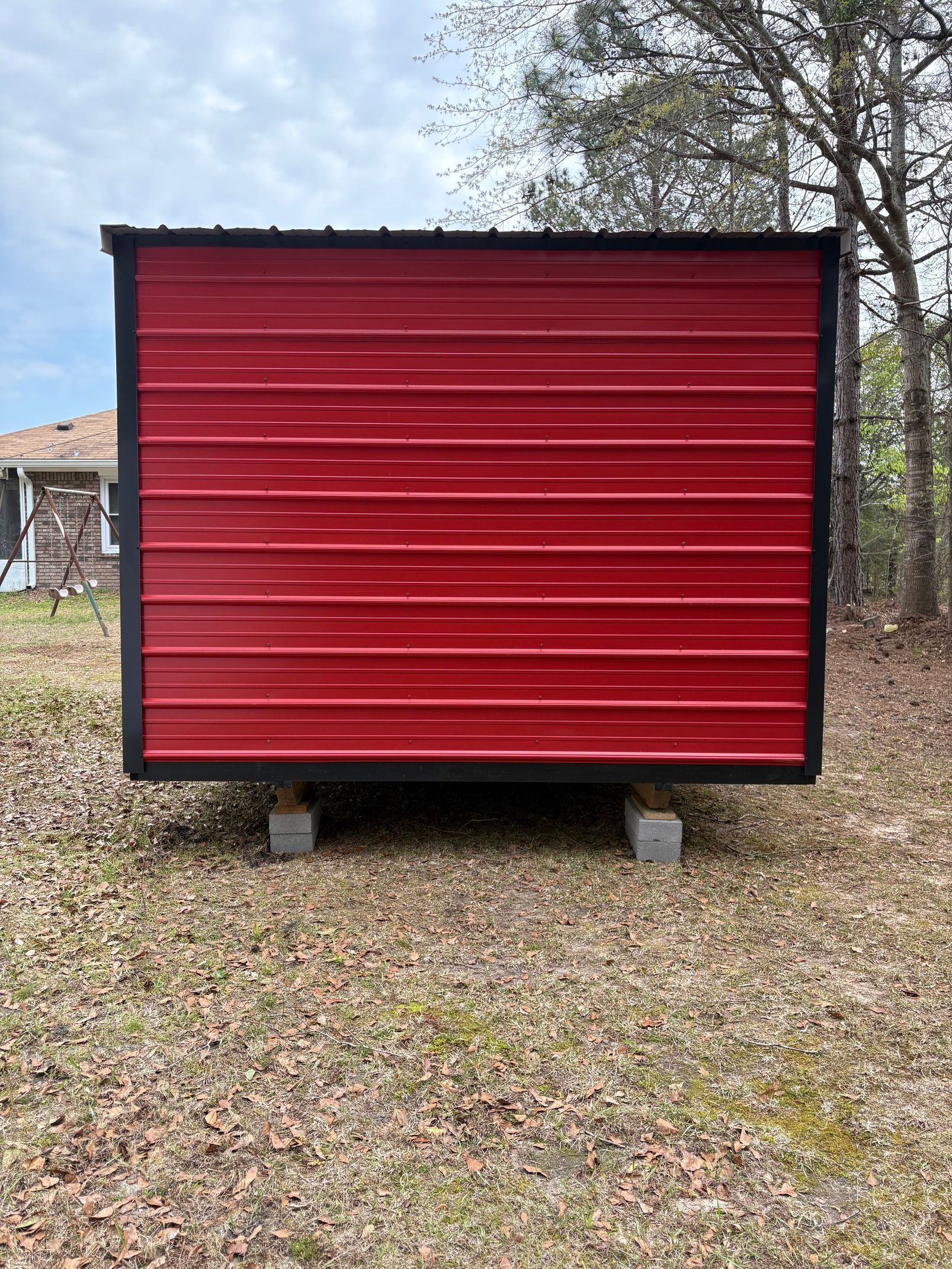 A rectangular, red metal shed with black trim sits on concrete blocks in a grassy yard.