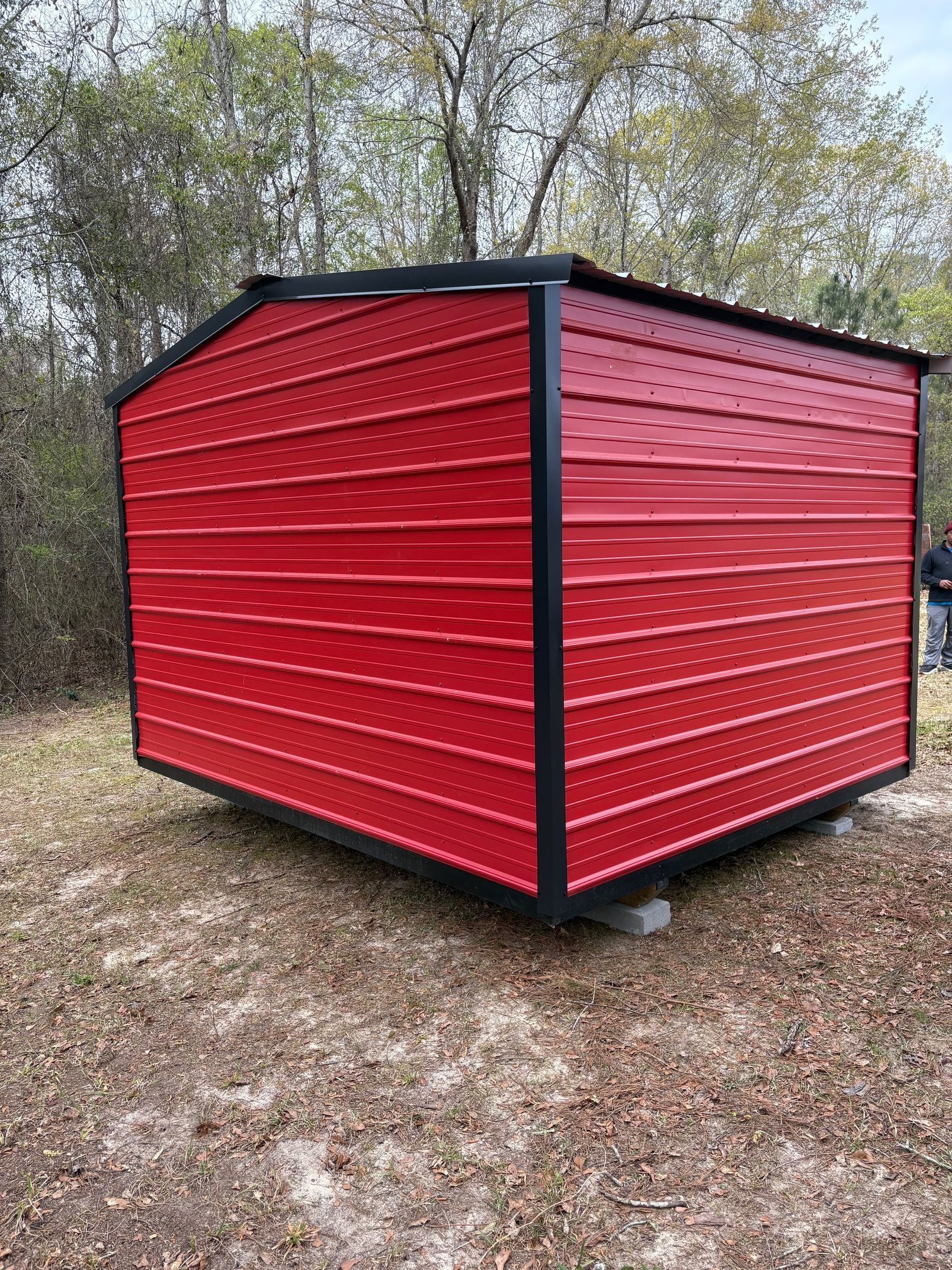 A red metal shed with a black frame sitting on grass in a wooded area.