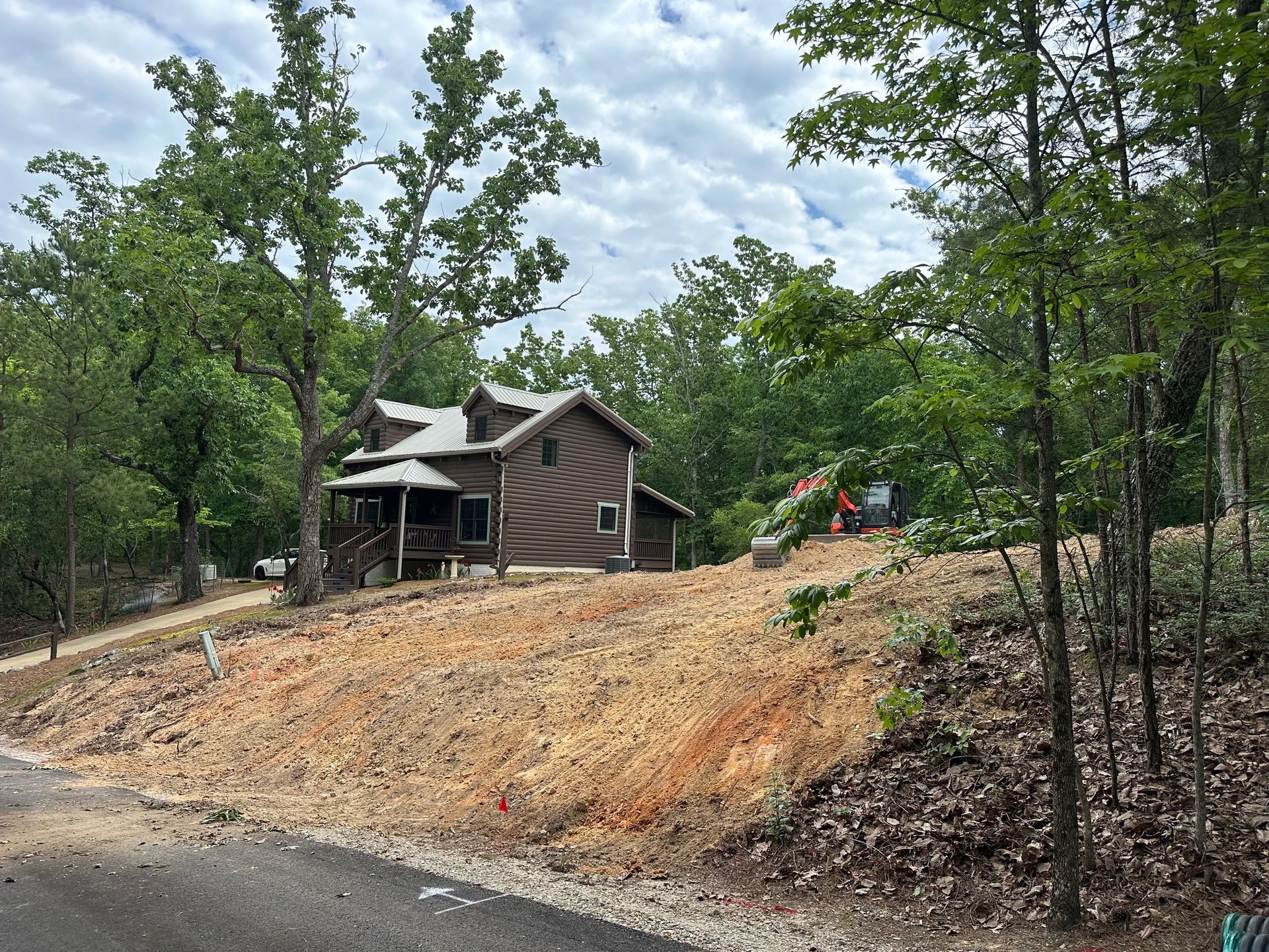 House on a cleared wooded hillside beside a dirt road under a cloudy sky