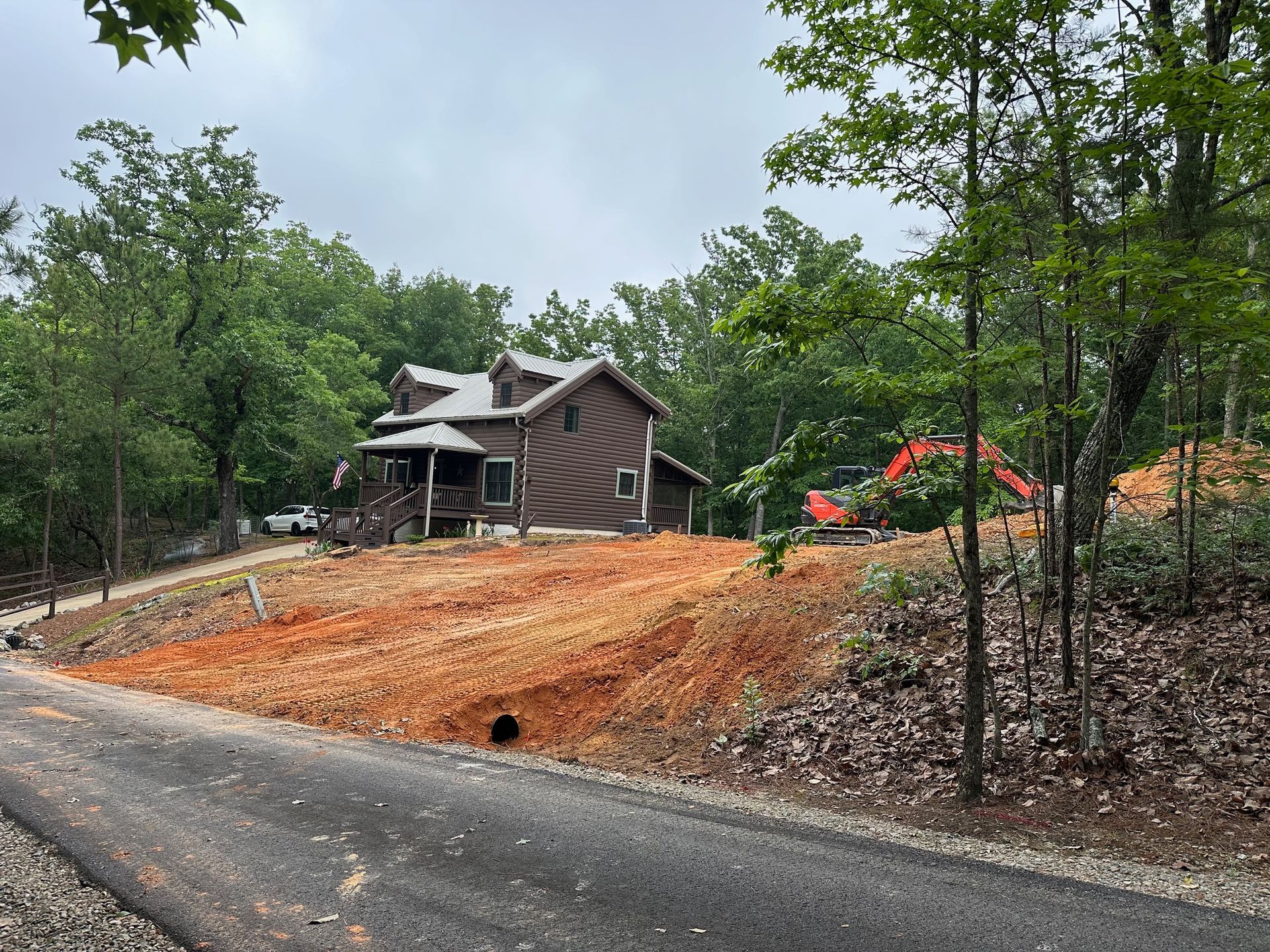 Rural dirt road beside a wooden house and trees, with orange construction soil on a wooded hillside