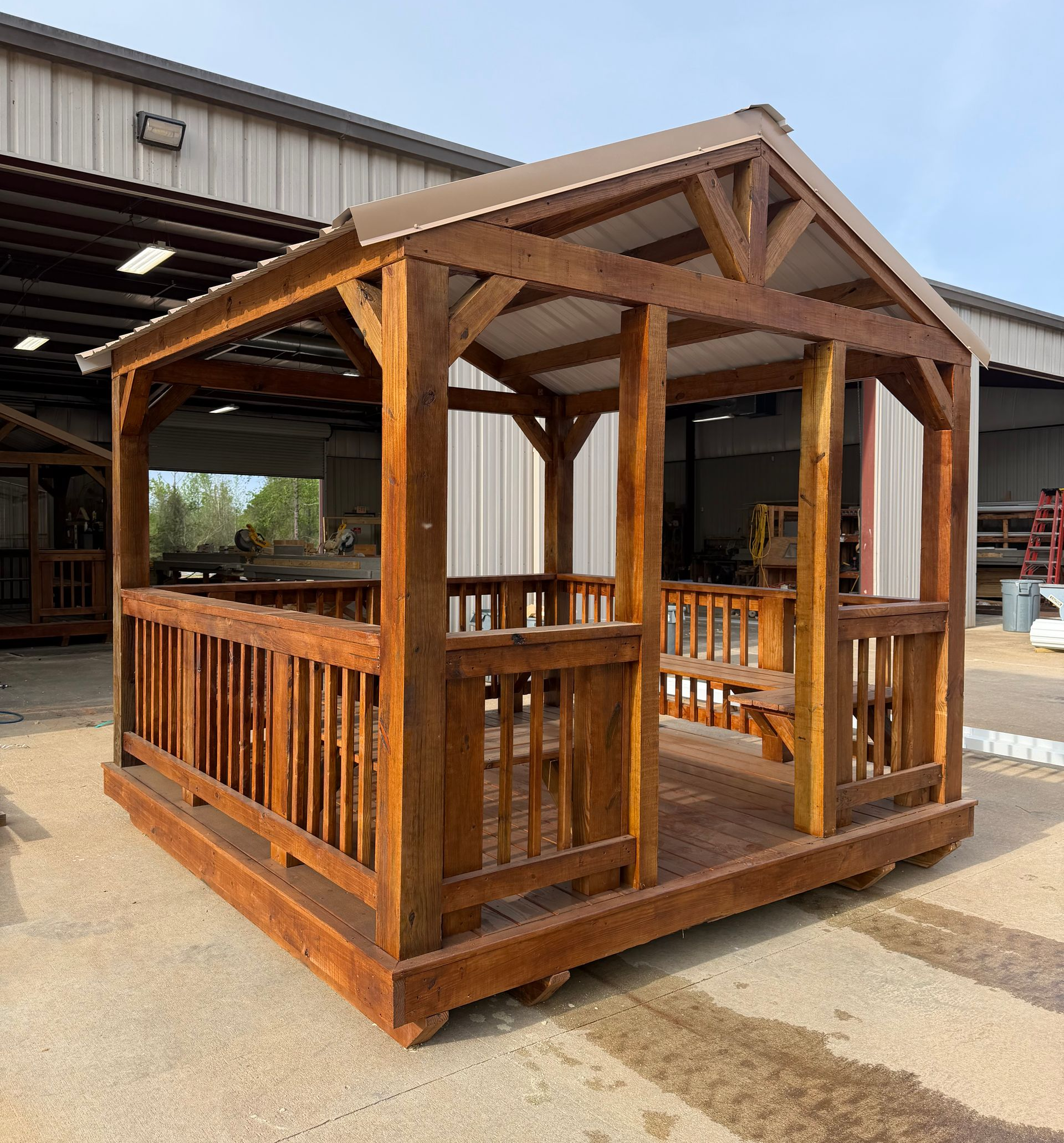 A wooden gazebo with a metal roof and railings sits on a paved lot in front of a warehouse.