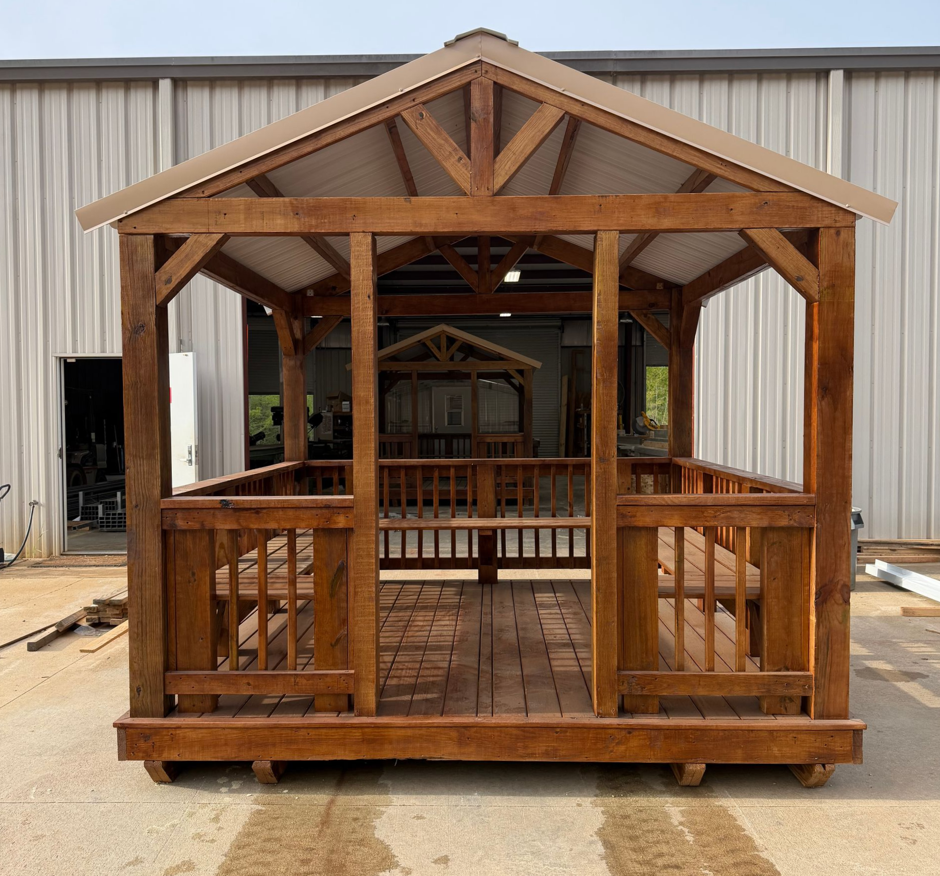 A wooden gazebo with a gabled roof and built-in benches, sitting on a concrete surface in front of a metal warehouse.