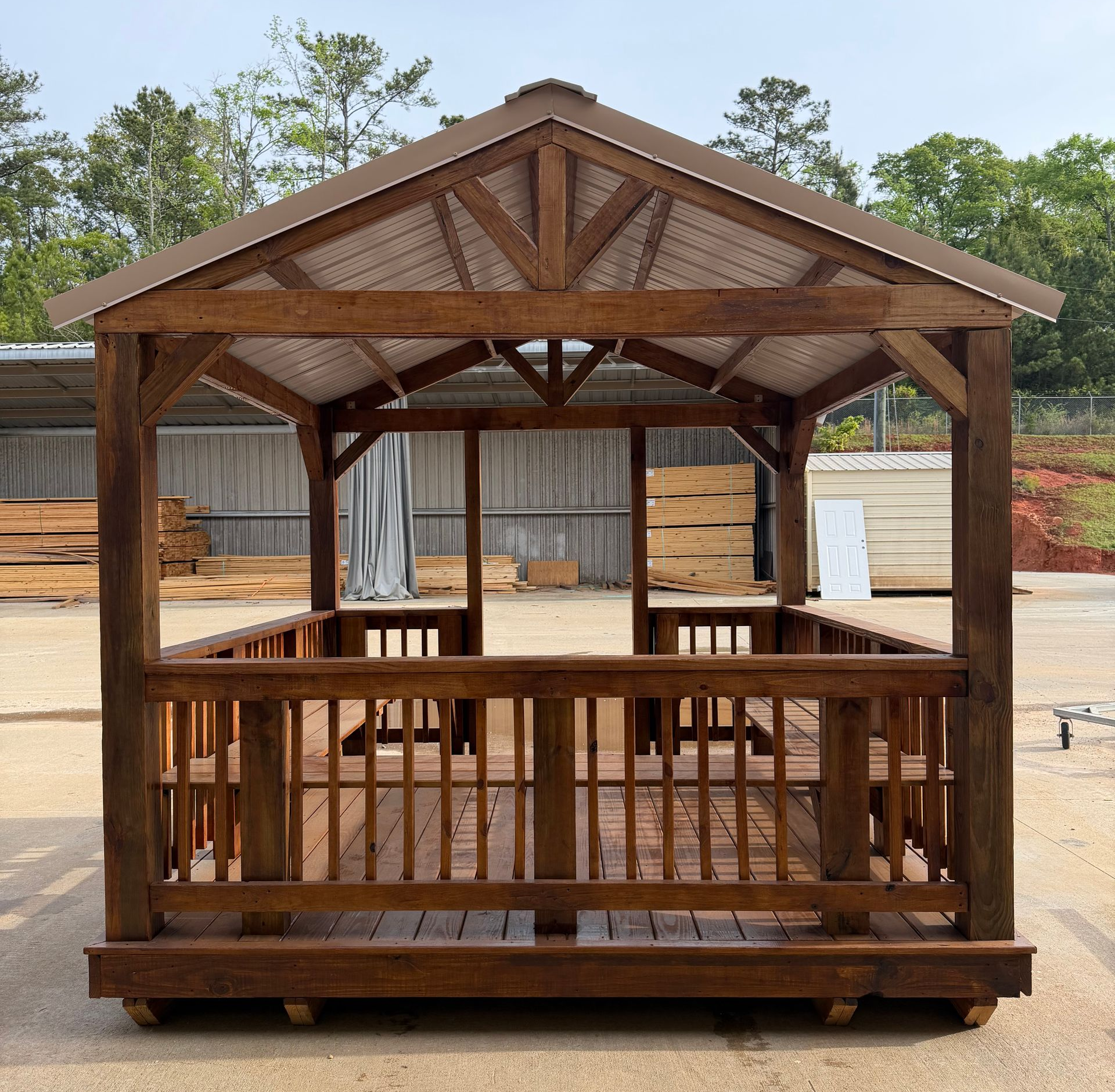 A wooden gazebo with a gabled roof and railings, centered on a paved outdoor lot with building materials in the background.