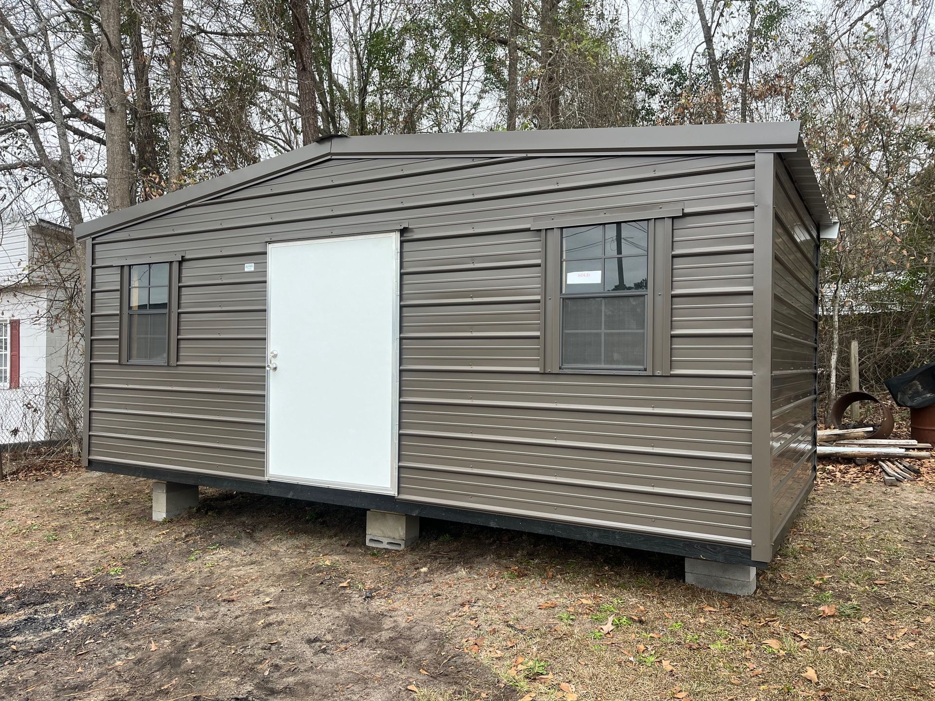 Brown shed with white door, two windows, set on concrete blocks, outdoors.