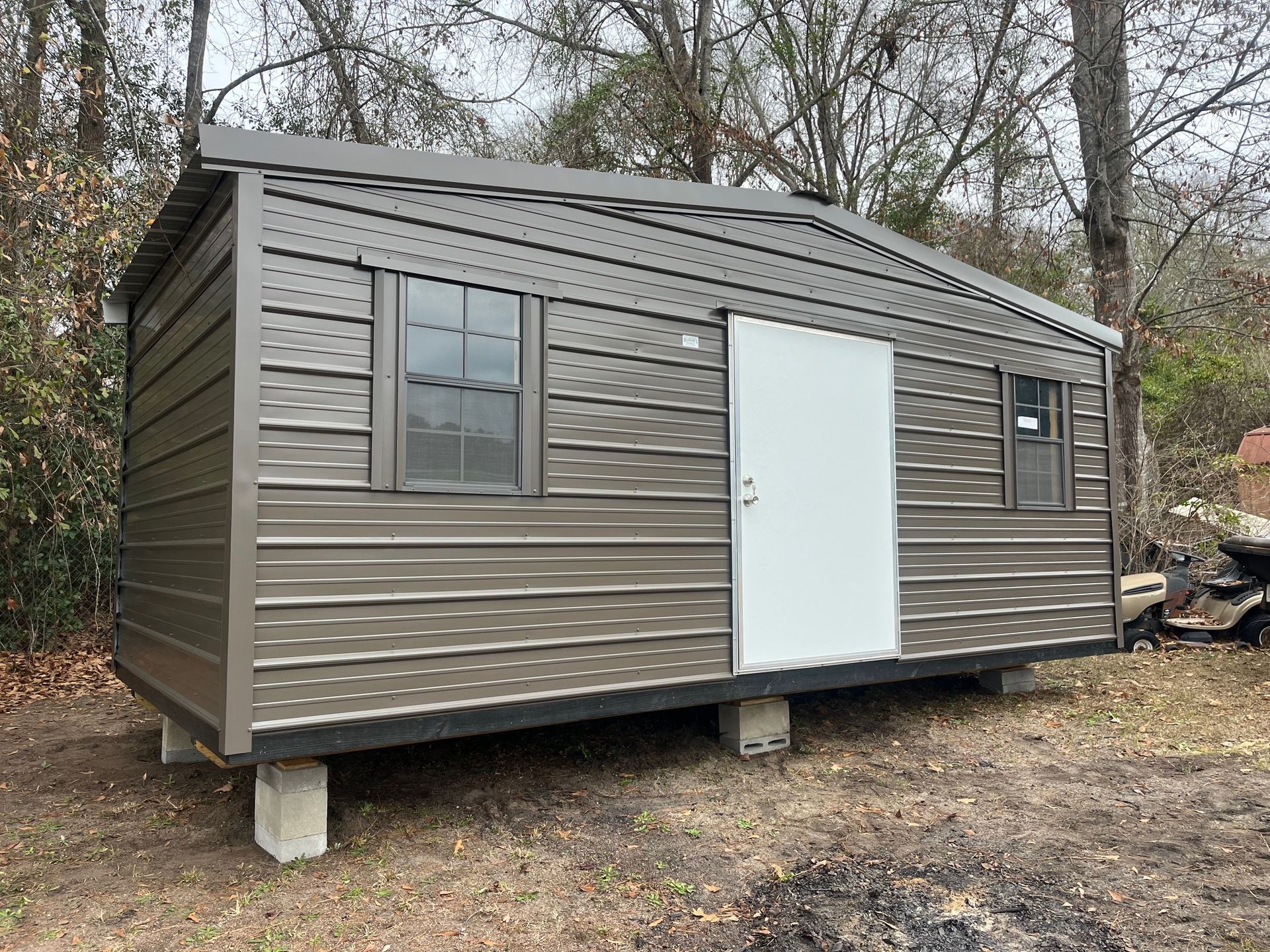 A brown metal shed with a white door and two windows on concrete blocks in a wooded area.