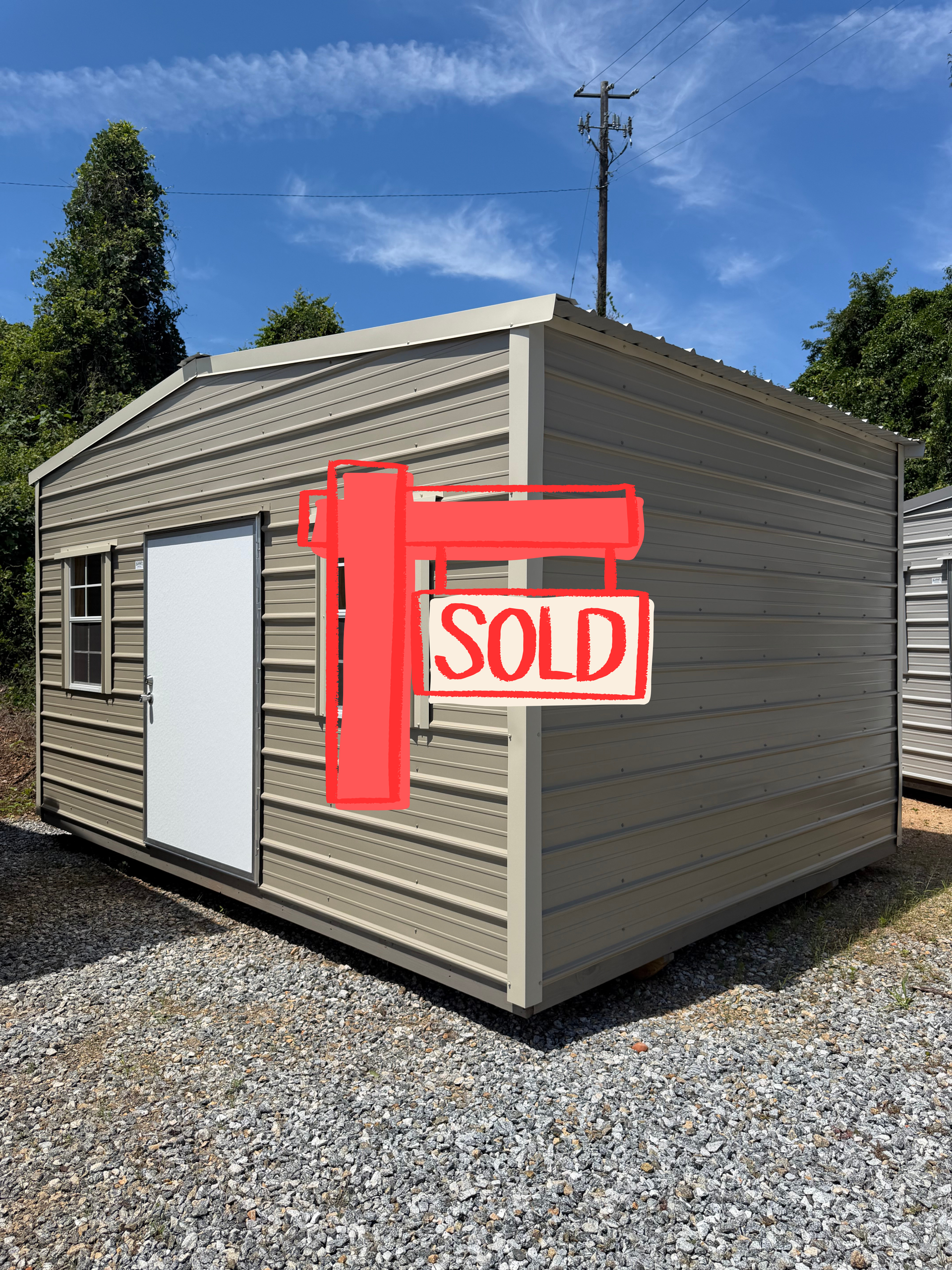 Small gray shed with a red “SOLD” sign on the side, set on gravel under a blue sky.