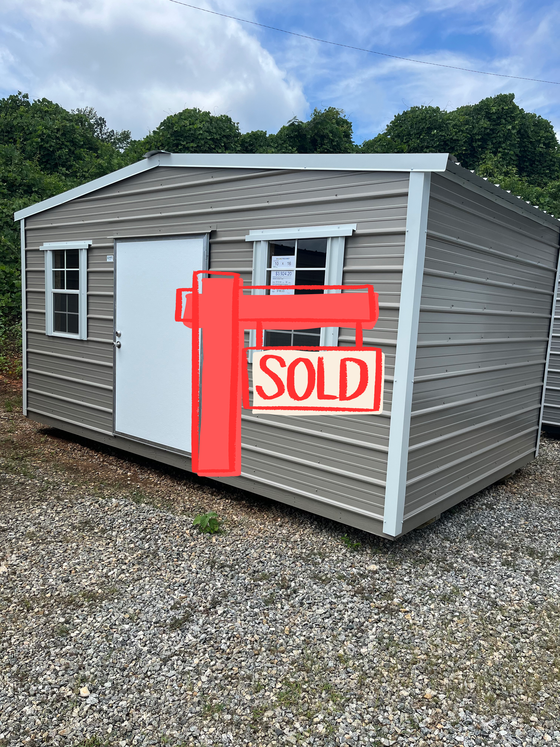 Small gray shed with red SOLD sign on gravel, outdoors under cloudy sky.