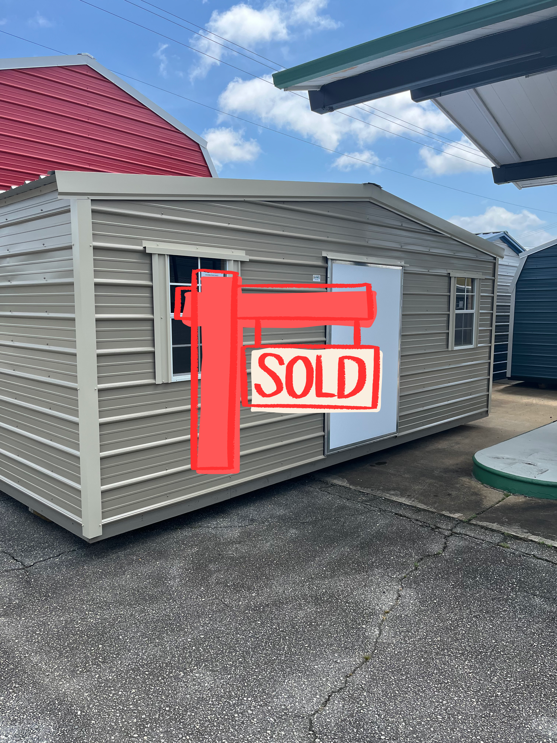 Small beige shed with a white door and window on a gravel lot under a blue sky.