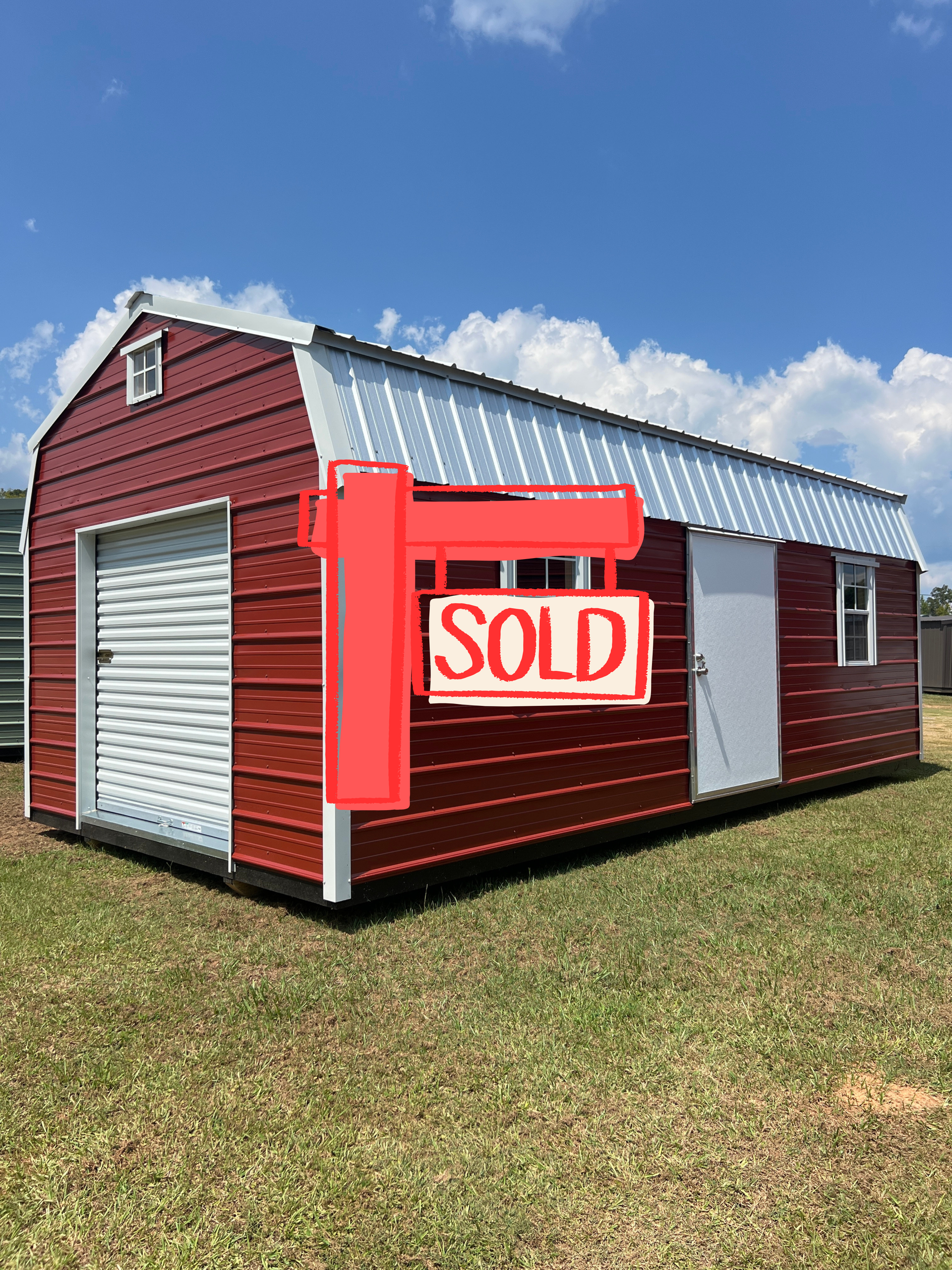 Red shed-style outbuilding with white trim, roll-up door, and a side window on a grassy lawn under blue sky