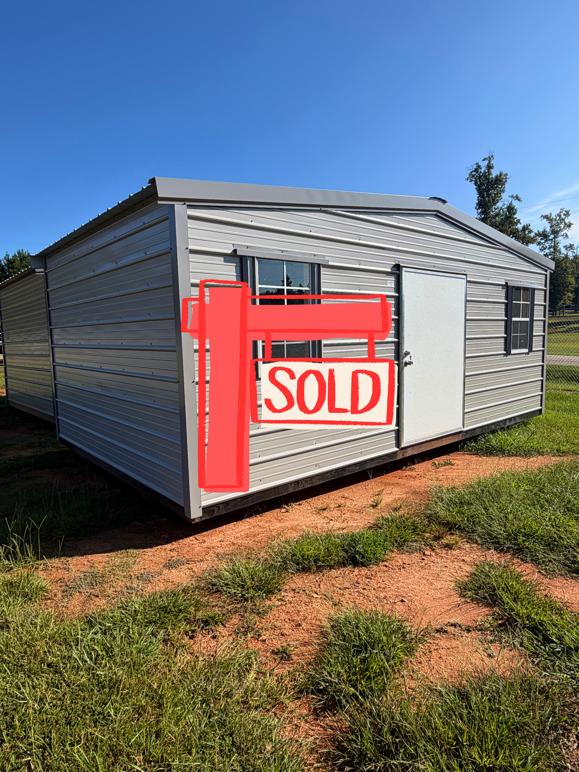Small gray metal shed with white door and windows on a sunny grassy lot