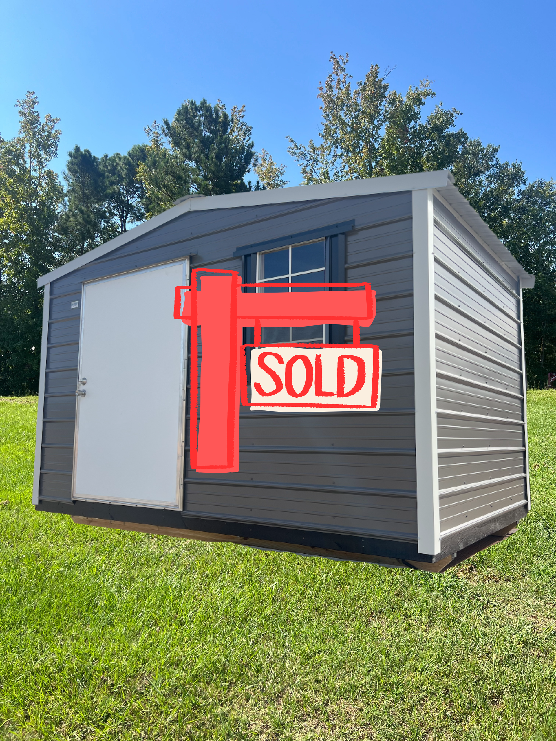 Small gray shed with white trim, a door and window, sitting on a grassy lawn under a blue sky.