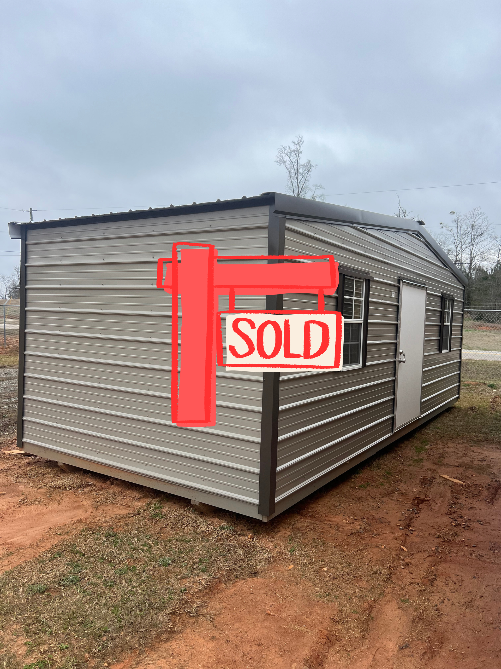Gray metal shed with a red “SOLD” sign on the side, sitting on a dirt lot under an overcast sky