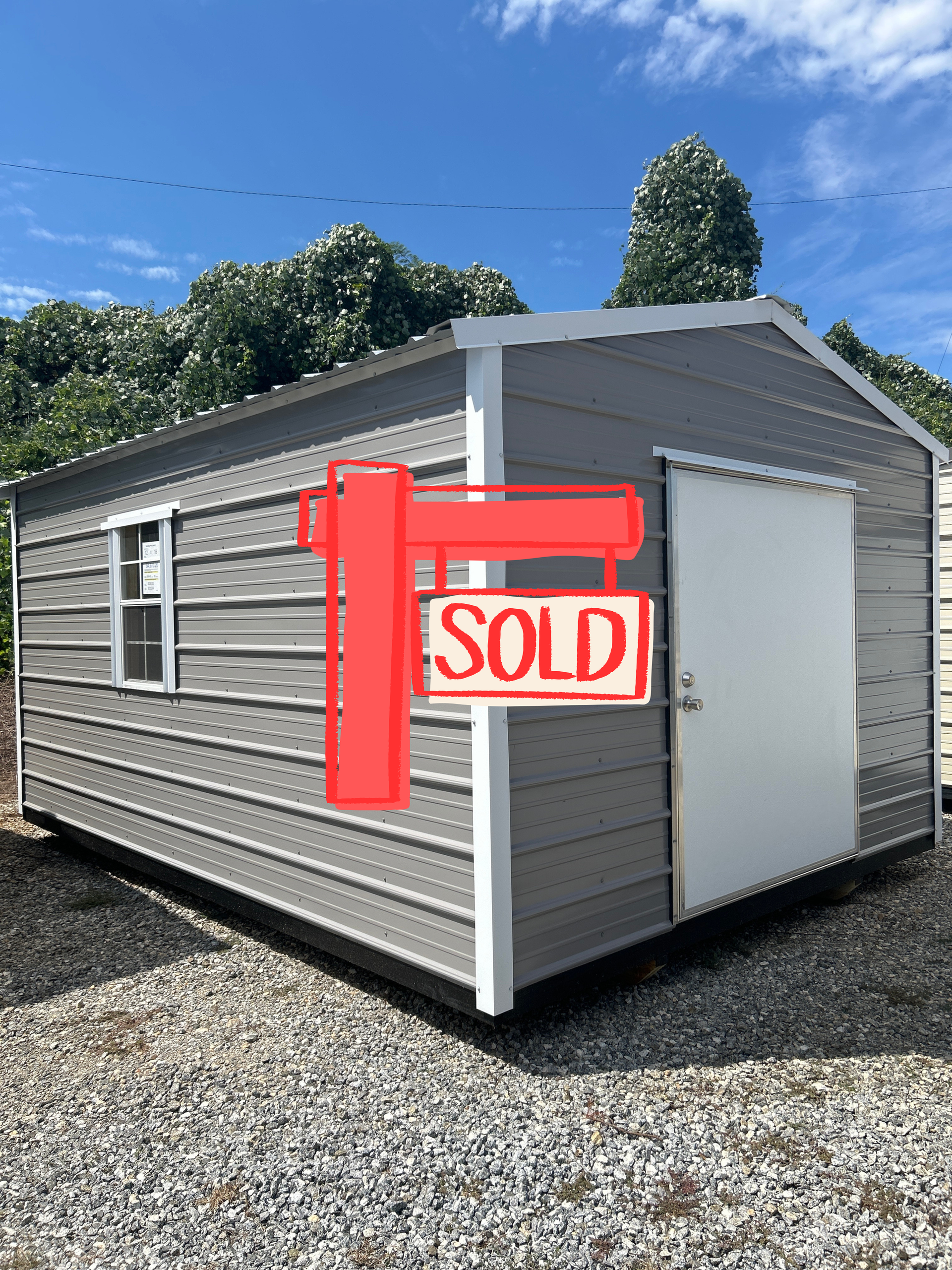 Gray modular shed with white trim and a red “SOLD” sign on a gravel lot