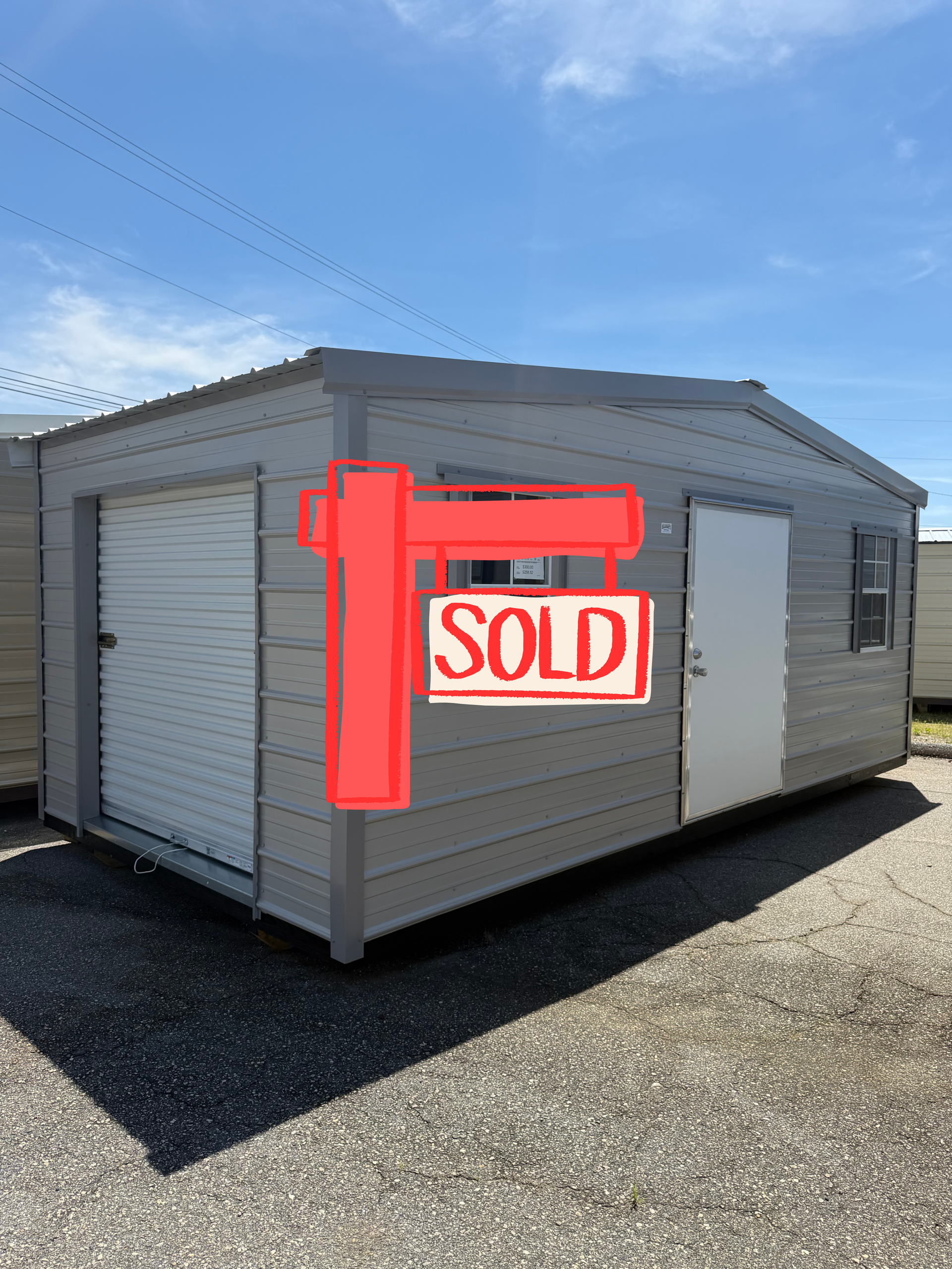 Gray portable shed with roll-up door and windows on a gravel lot under a blue sky