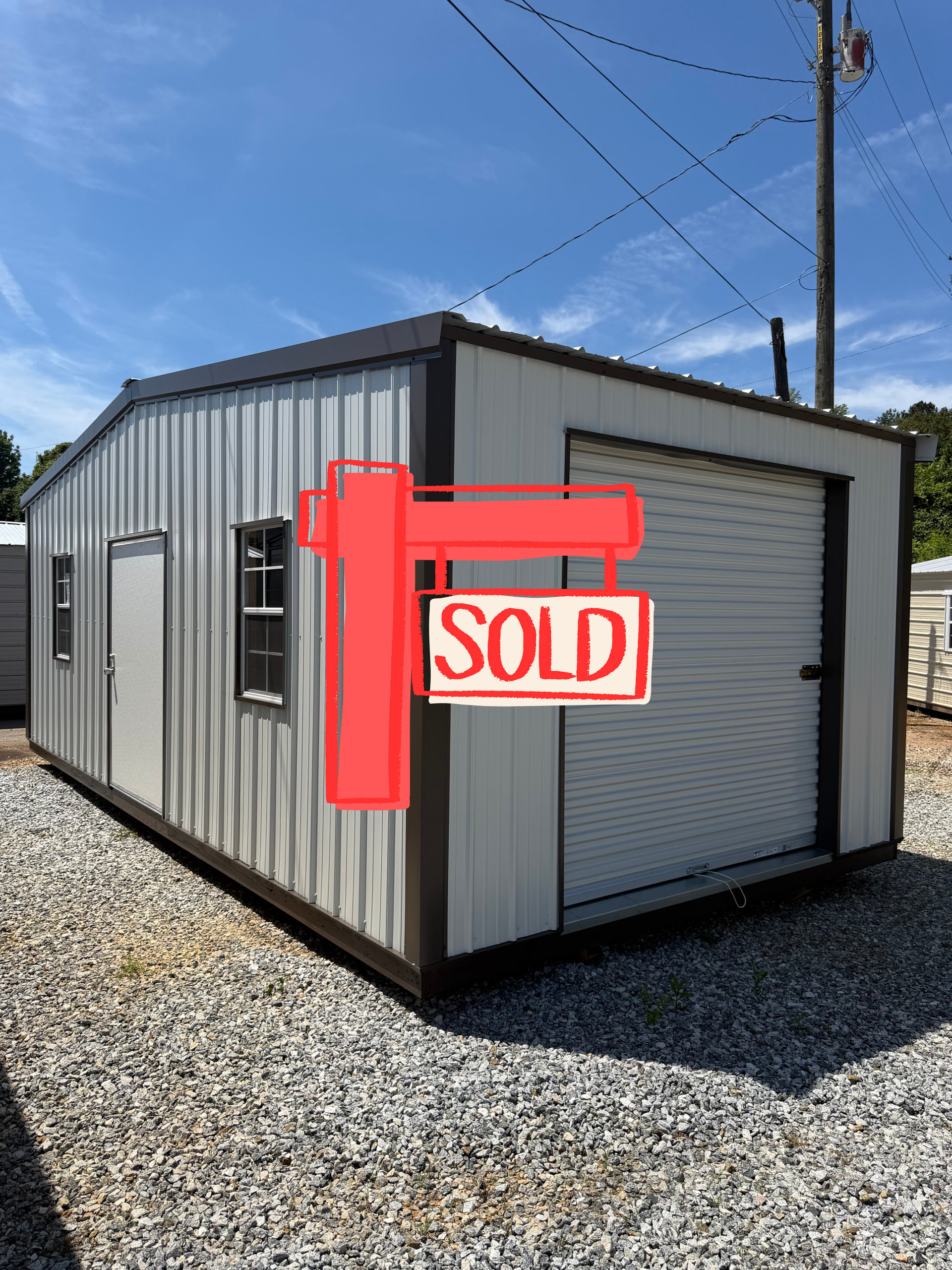 Gray storage shed with a white roll-up door, marked “SOLD” in red on a gravel lot.