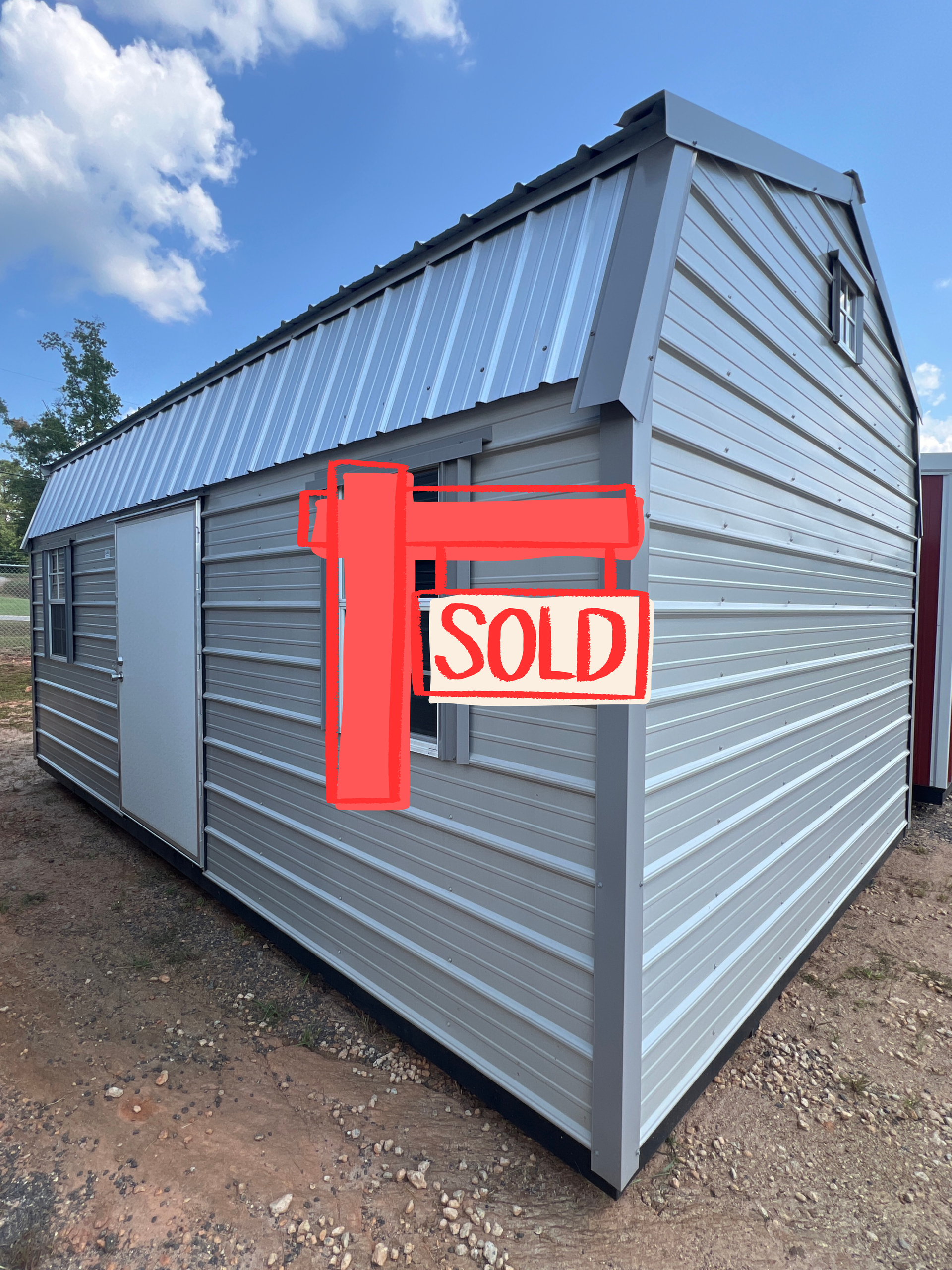 Gray metal shed with a red sold sign on the front, viewed from the corner outdoors.