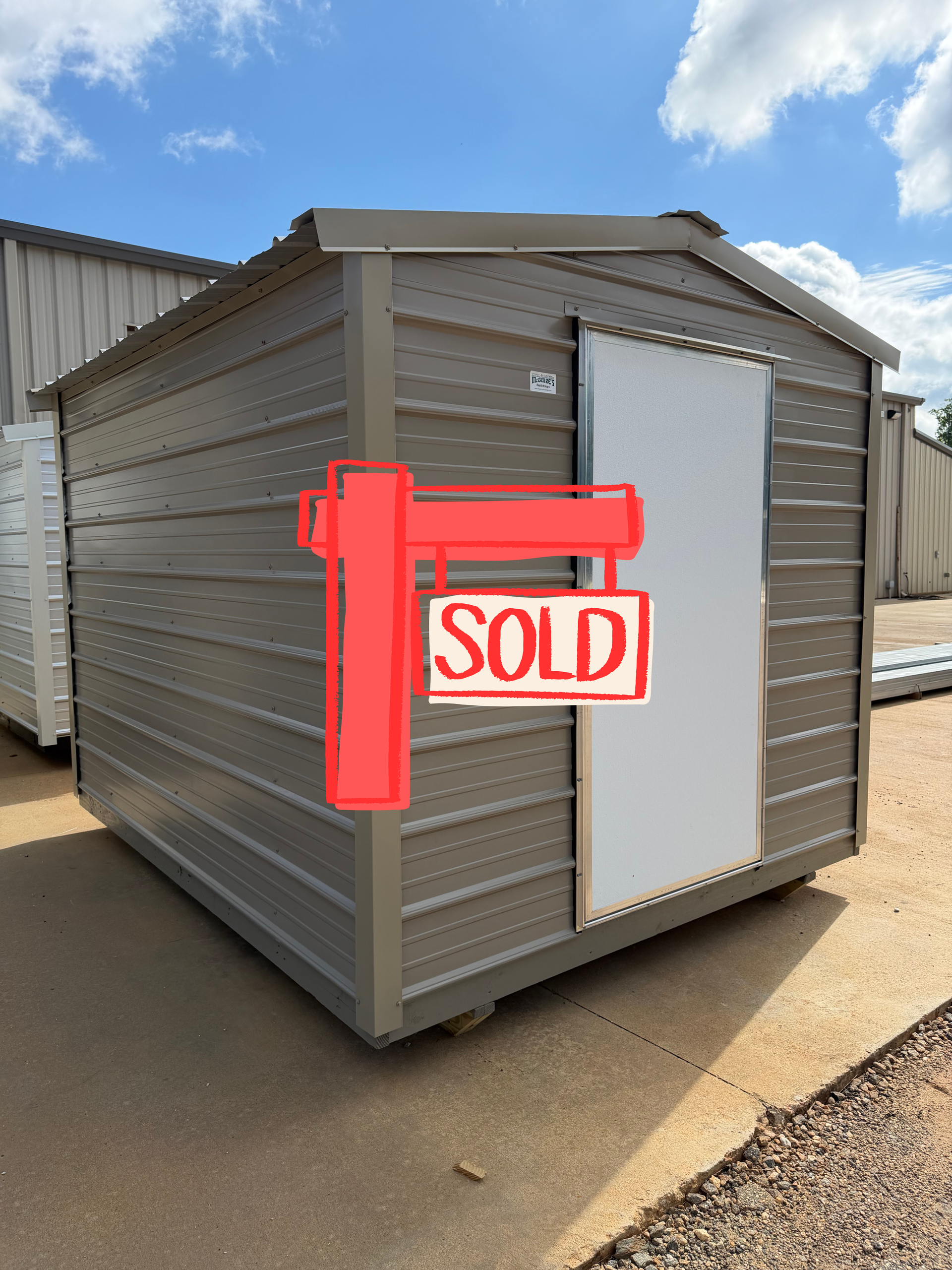 Small gray metal storage shed with a white door and a red “SOLD” sign on the front.