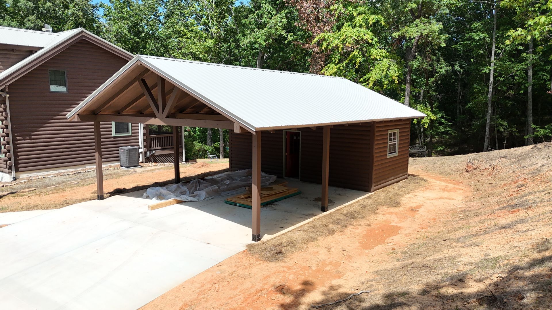 Detached carport beside a brown cabin, with a concrete driveway and white metal roof
