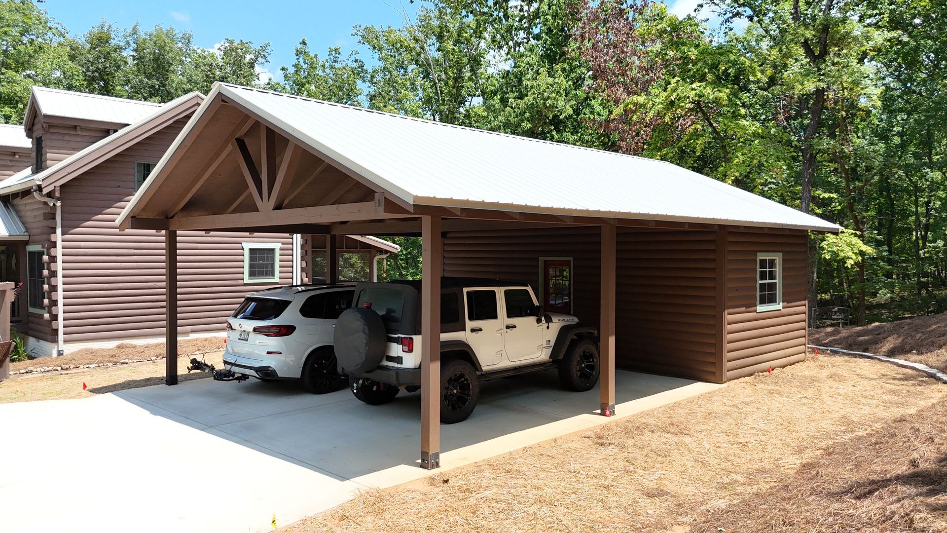 Two vehicles parked under a wooden carport beside a log cabin in a wooded area.