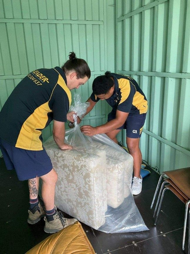 Two movers wrapping a beige couch in plastic inside a storage unit - — Leader Removals & Storage in Mitchell, ACT