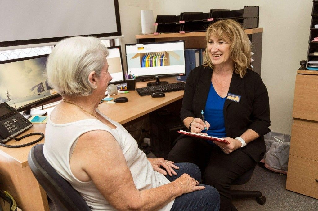 Woman in office setting speaking with another woman, both smiling. One holds clipboard. Desk with computer behind them.