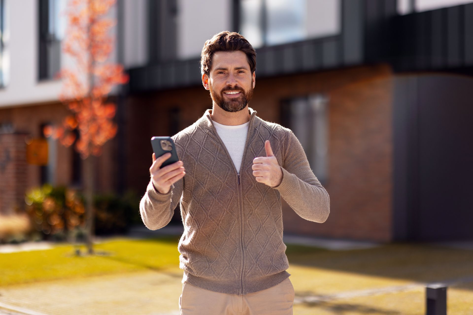 Man holding phone, giving thumbs up outside modern building, smiling.