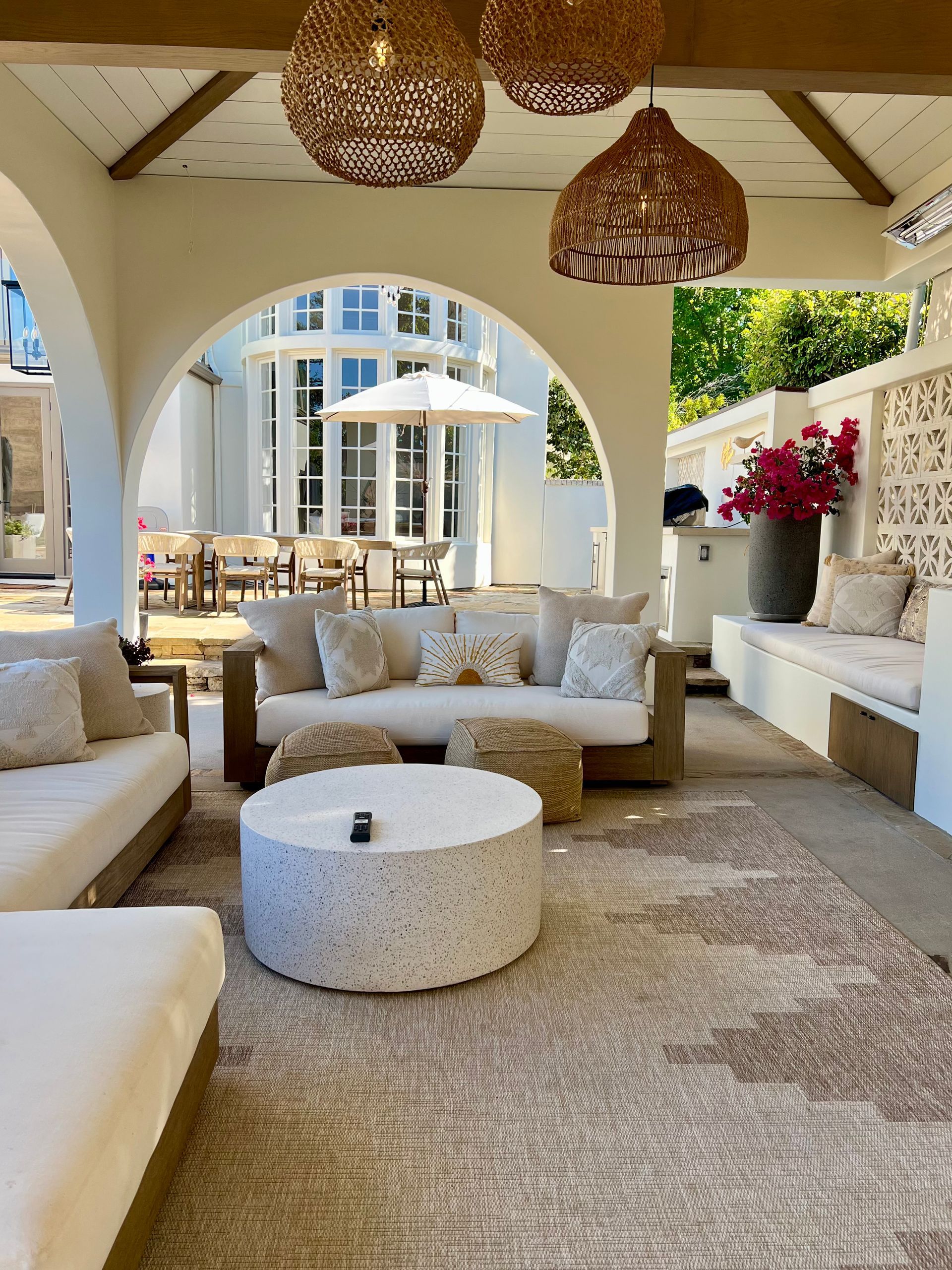 Covered outdoor patio with white cushioned seating, woven lamps, and a central round table overlooking a pool and buildings