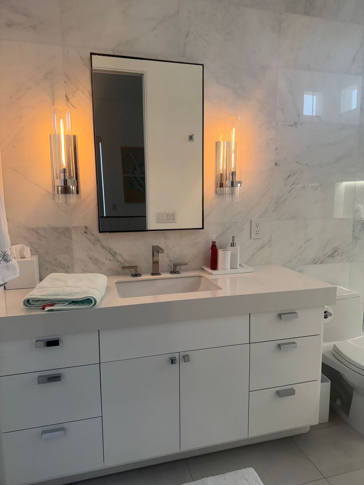 White bathroom with marble walls, white vanity, silver fixtures, and rectangular mirror. Two sconces flank the mirror.