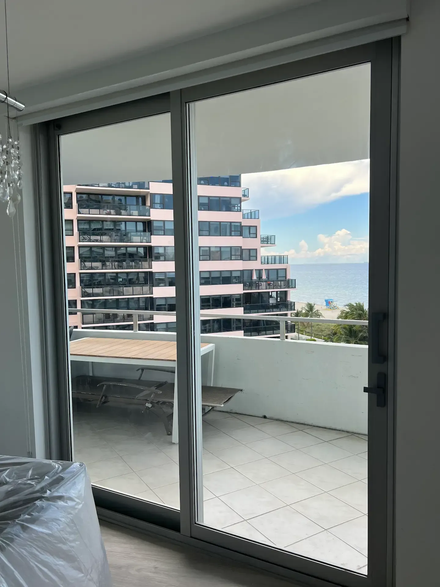 Sliding glass doors open to a balcony with white tile, overlooking modern buildings and the sea.