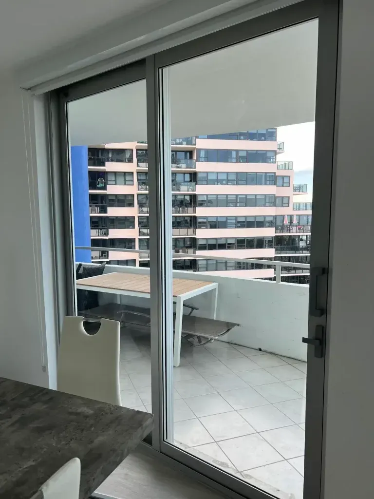 Sliding glass door to a balcony with a table and chairs, overlooking apartment buildings outside