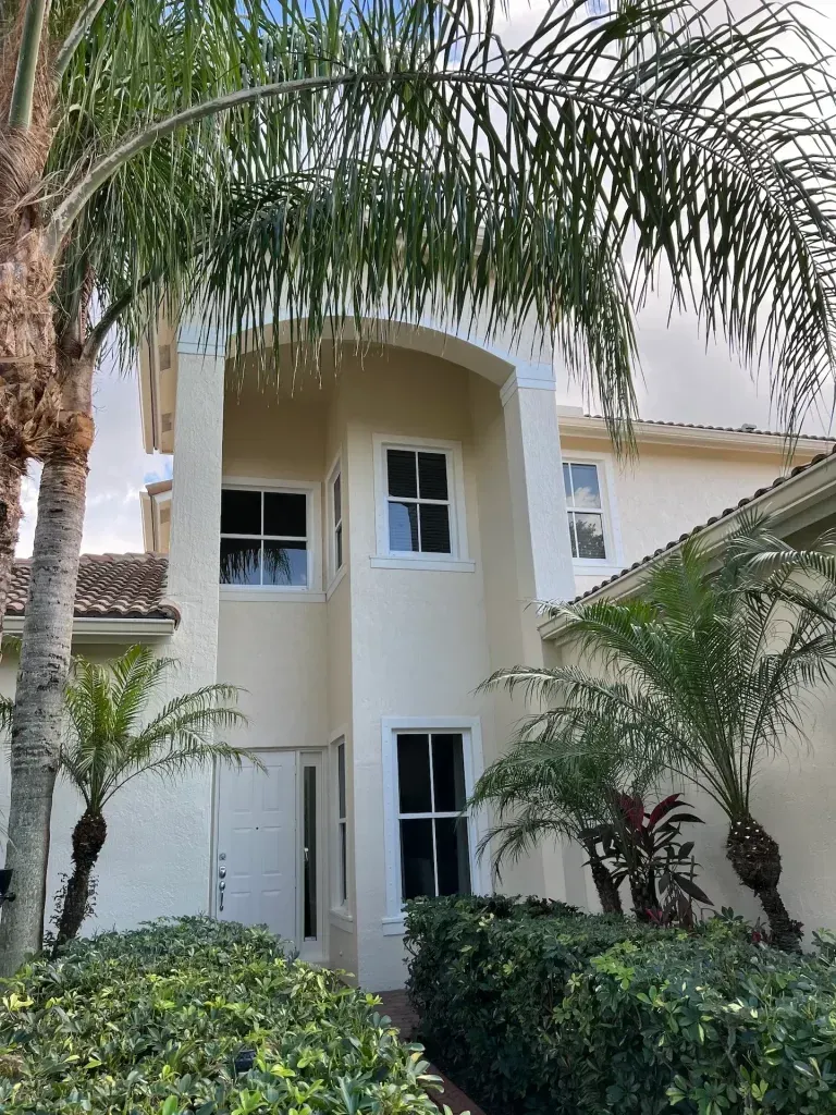 Two-story white house with arched entry, framed by palm trees and dense green shrubs.