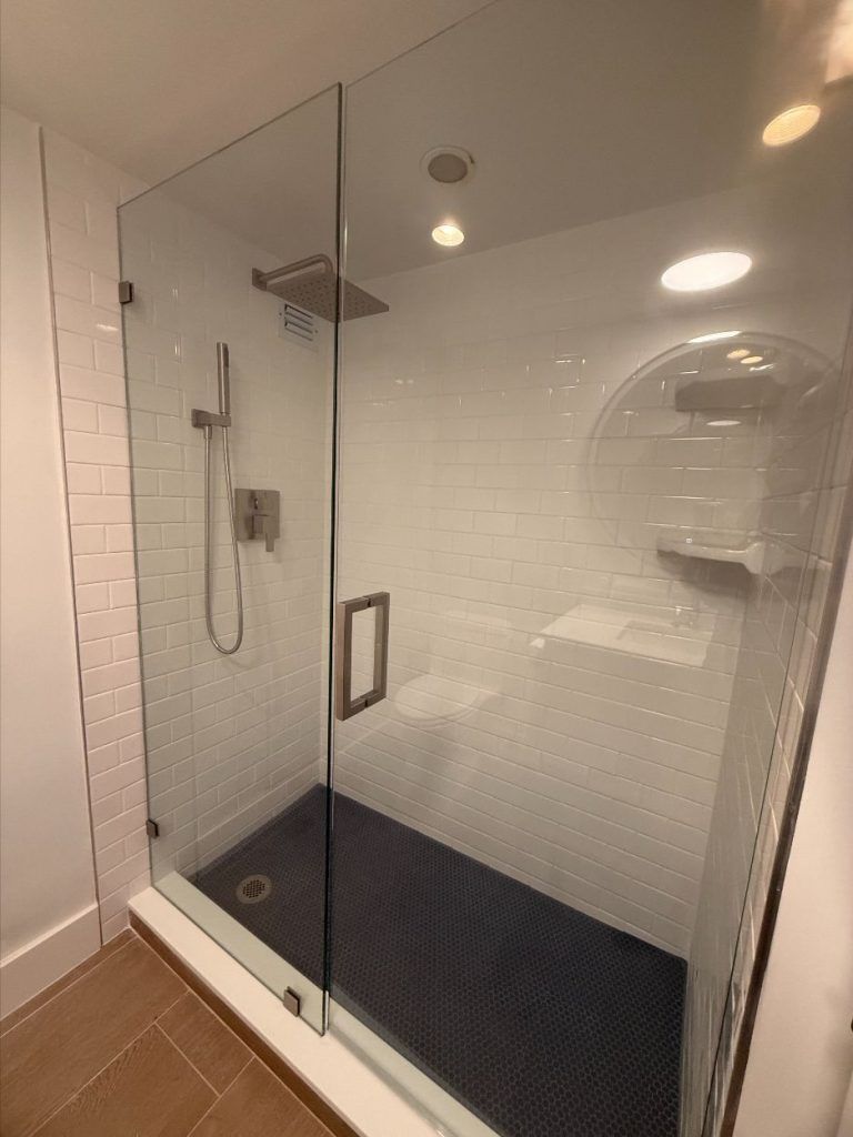 Glass walk-in shower with black floor mat, wall-mounted showerhead, and white tile bathroom walls