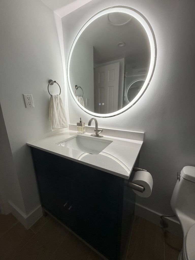 Small white bathroom vanity with round backlit mirror and sink, next to a toilet.