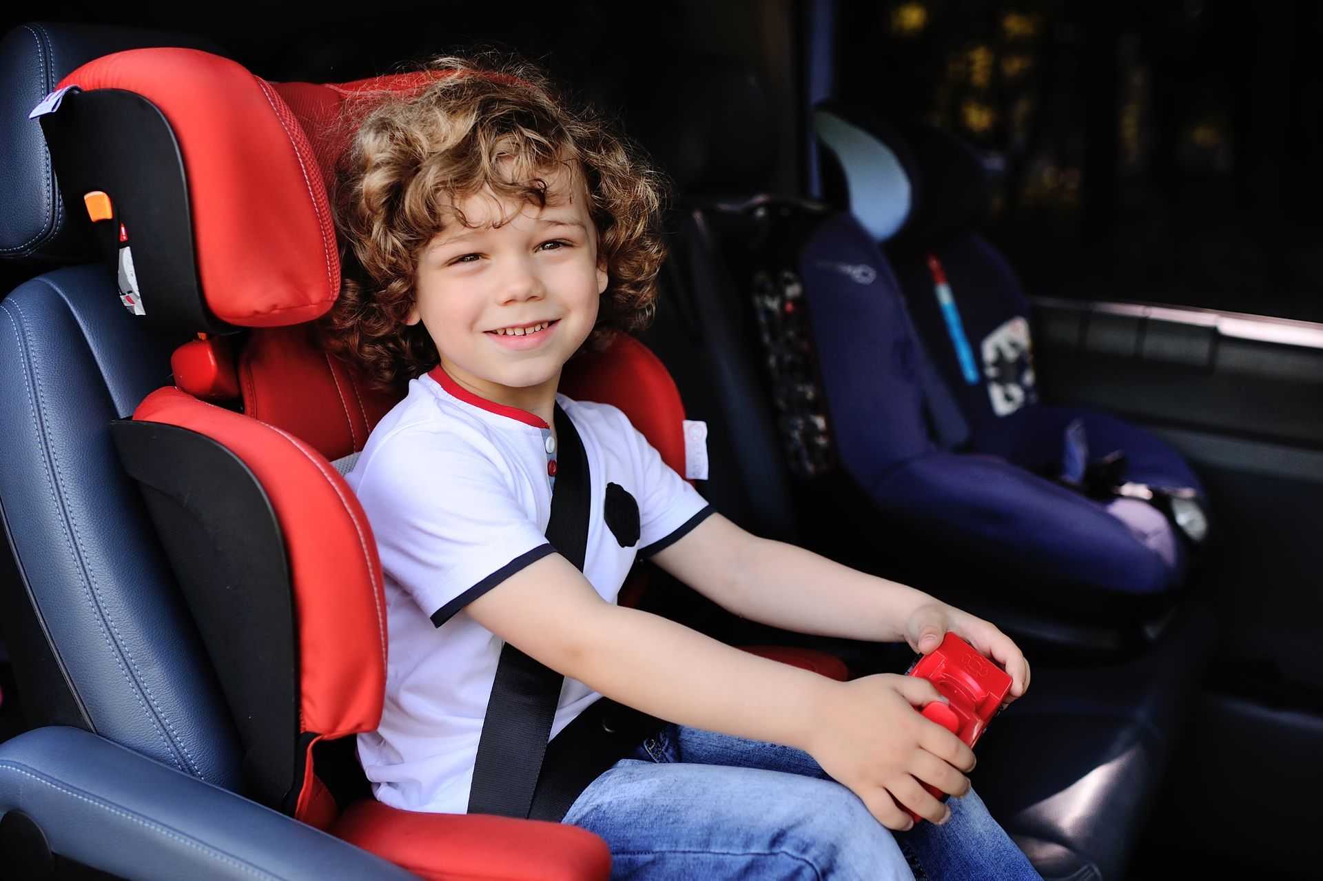A curly-haired child with a cheerful expression sits in a red and black car seat inside a vehicle.