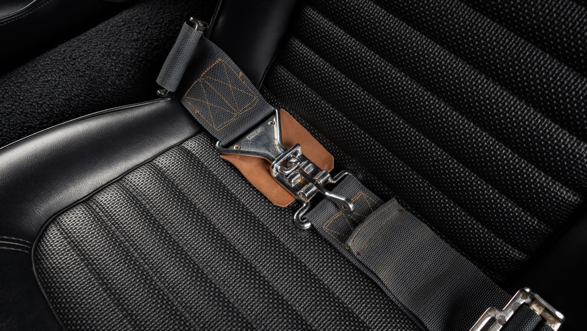 A close-up of a black, multi-point seatbelt buckle fastened over a textured black car seat with a tan accent piece.