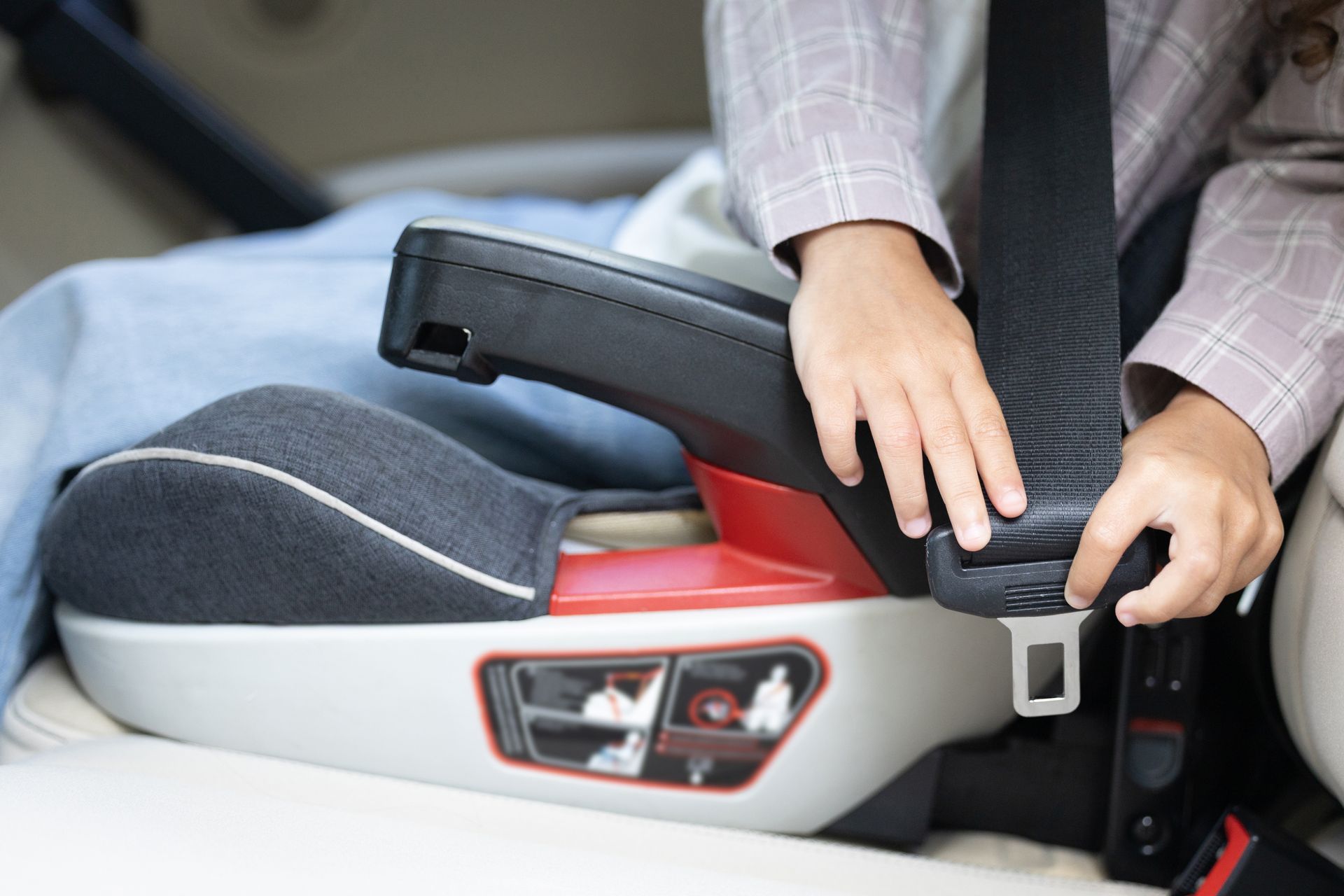 A person placing a vehicle seat belt into the guide slot of a gray and red booster seat.