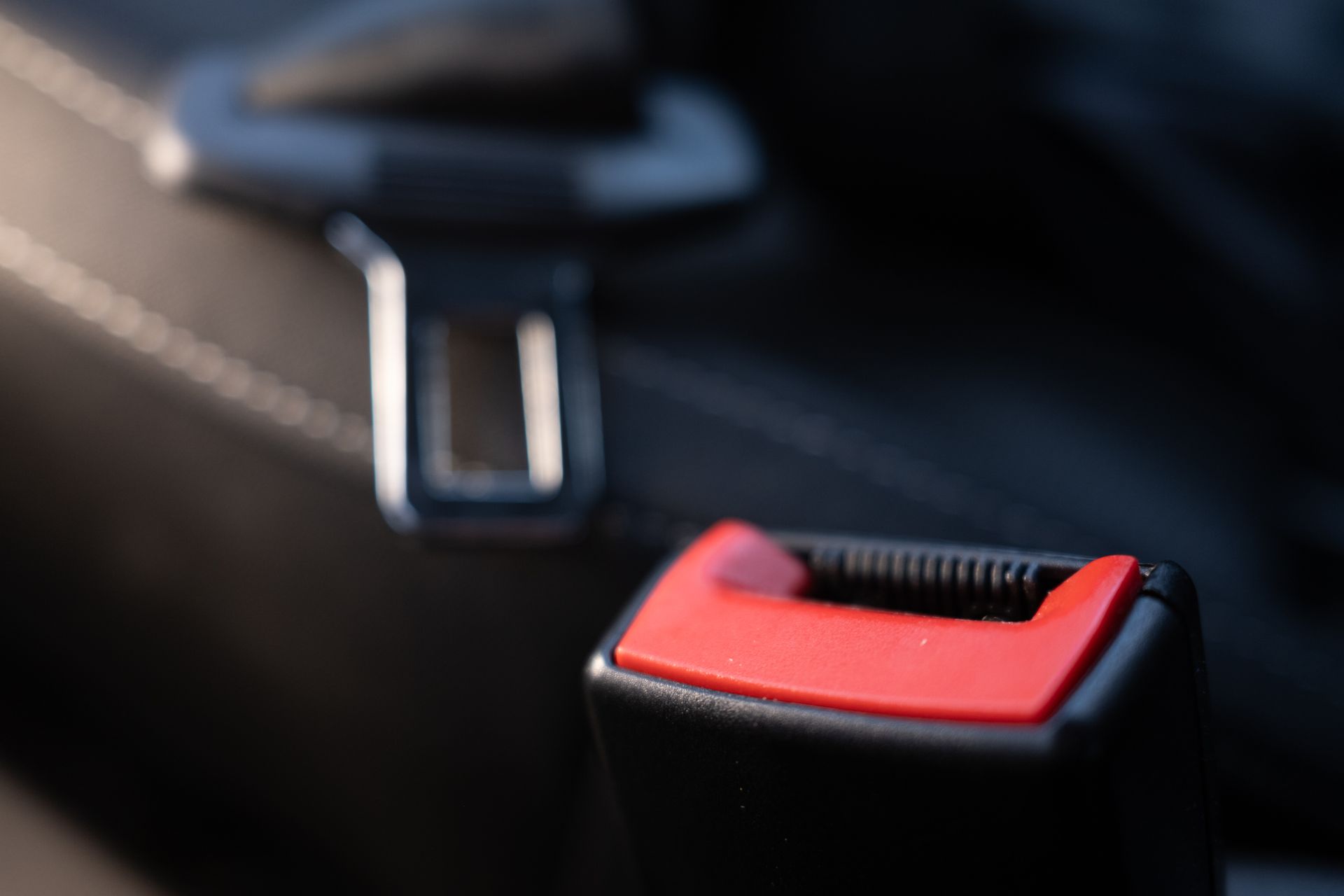 A close-up of a black car seat buckle with a red release button and an out-of-focus seat belt latch in the background.