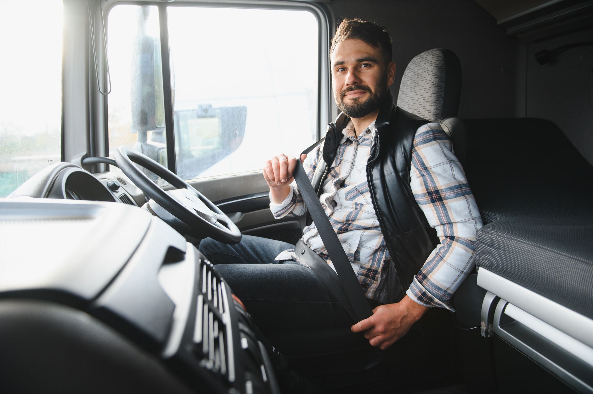 A person in a plaid shirt and vest sits in the driver's seat of a truck, fastening their seatbelt while looking at camera.